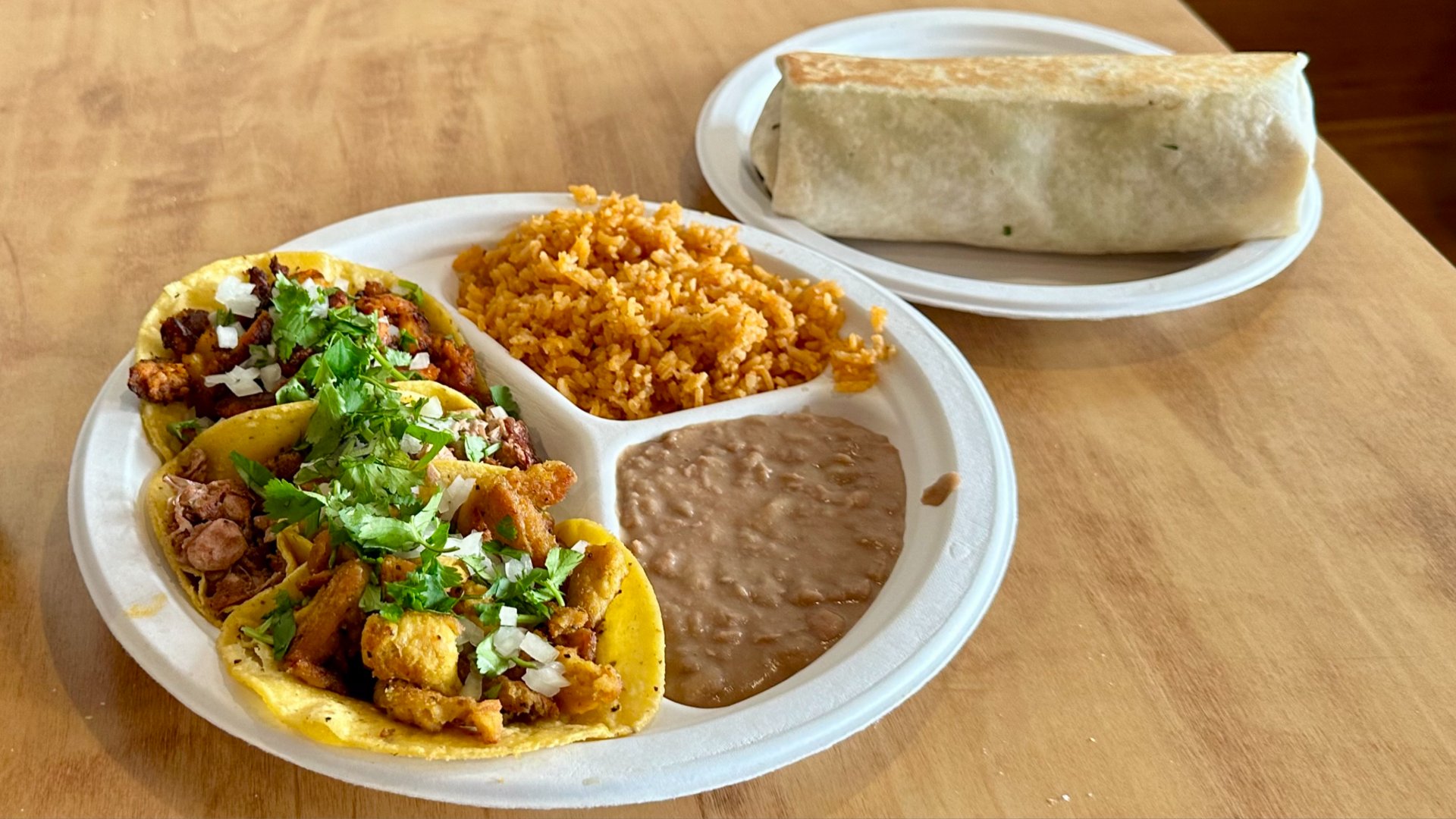 Three tacos, rice and beans on a plate next to a plate with a burrito on a wooden table.