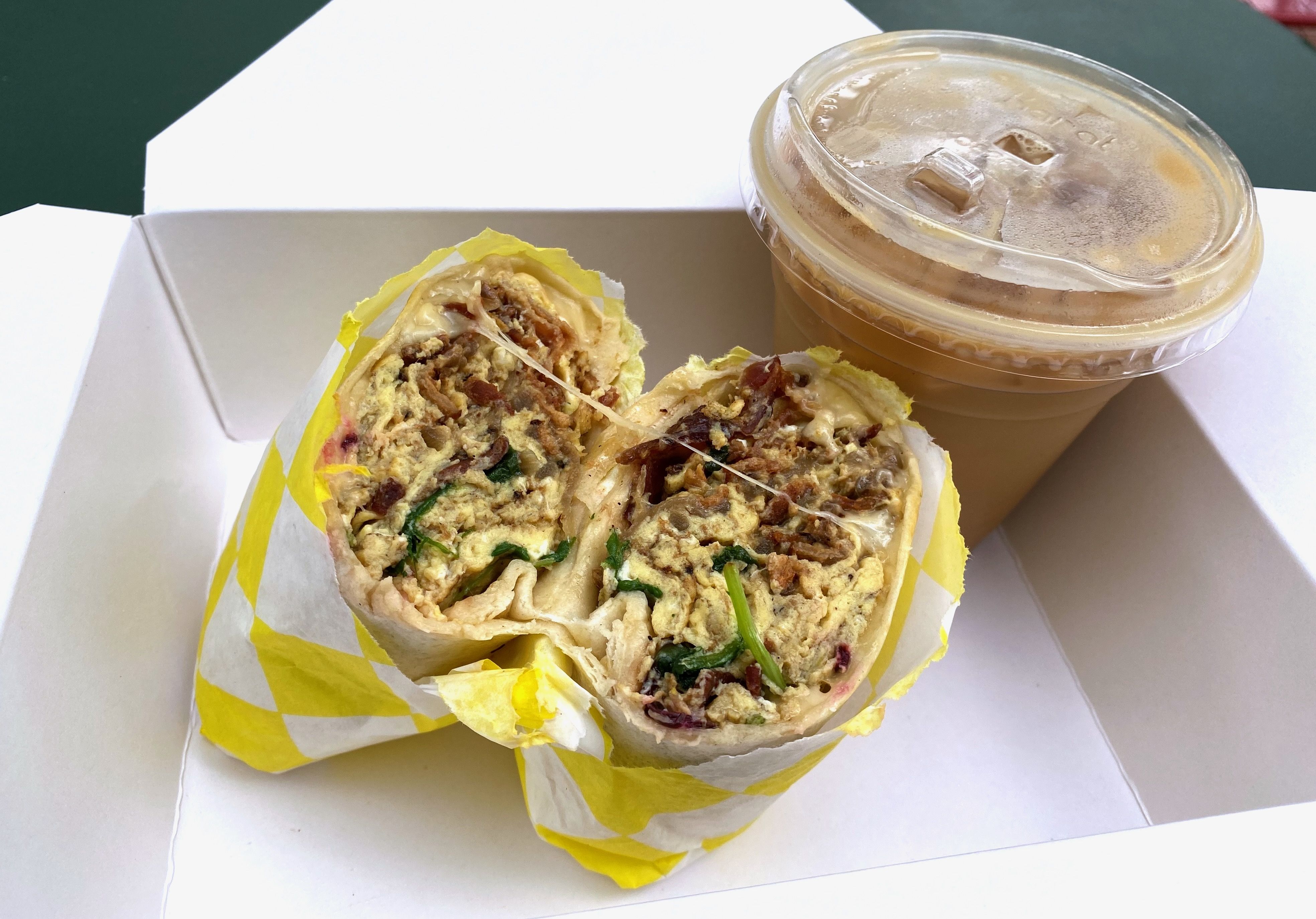 A breakfast burrito wrapped in yellow and white paper, cut in half, and an iced latte in a plastic cup