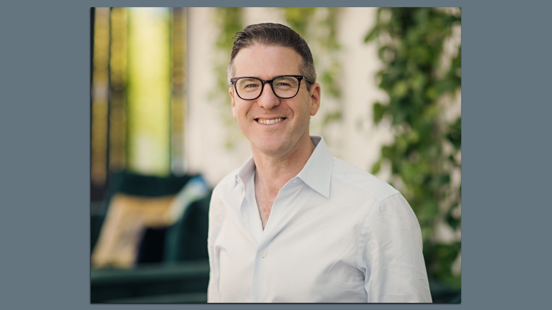 Portrait of a smiling man with dark hair and black-framed glasses, wearing a light button-down shirt, with blurred green plants in the background.