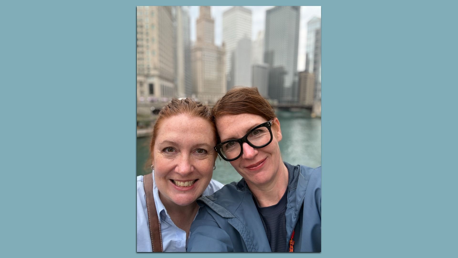 Two women smile for a selfie by a river with a blurred city skyline behind them. The woman on the right wears black glasses and a blue jacket; the other has red hair and a light blue shirt.