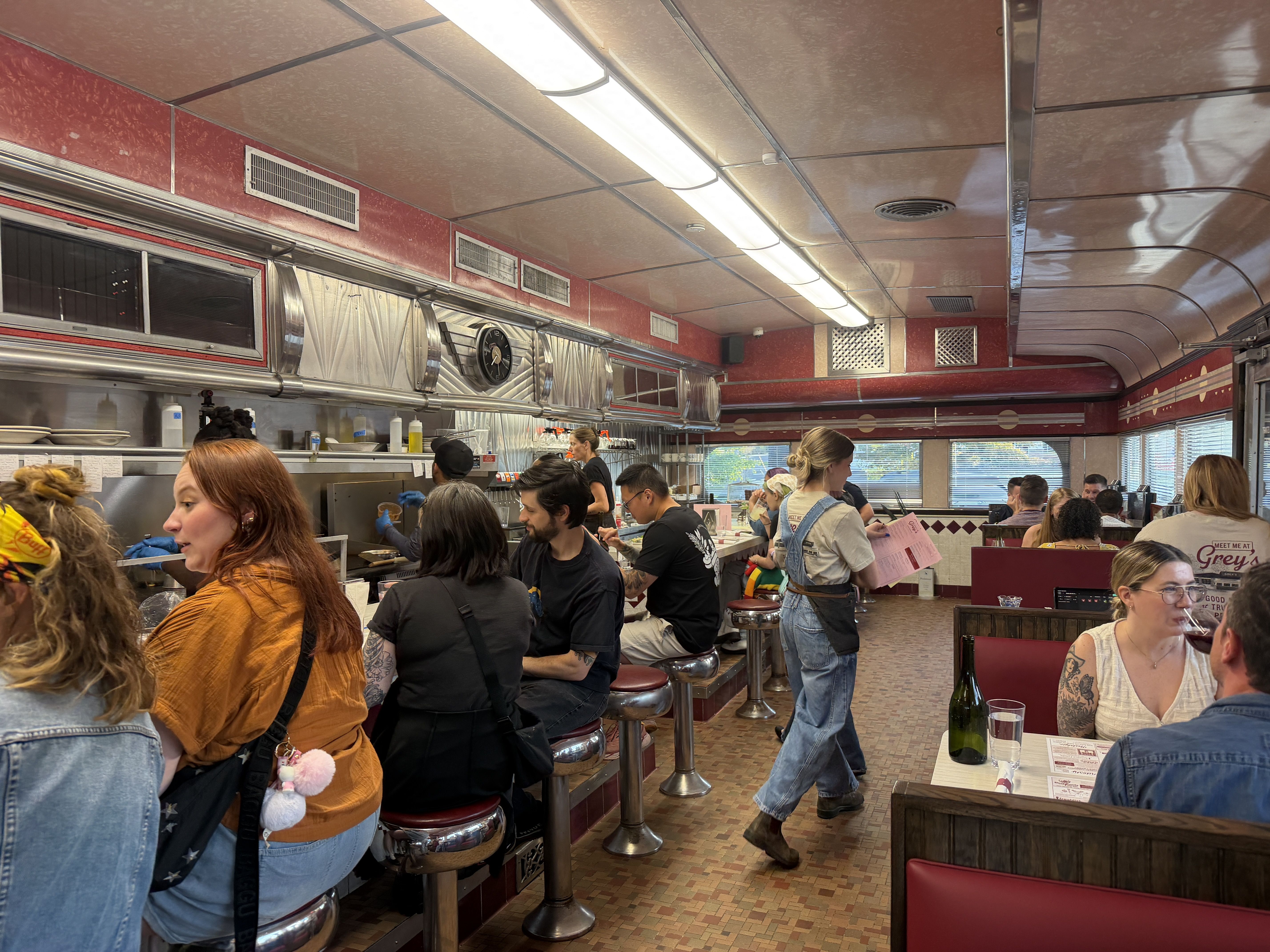 Busy retro diner scene: red walls, stainless steel kitchen, row of chrome stools along a counter, patrons ordering and eating. Bright overhead lights, curved metallic ceiling, a wine bottle on a front table.
