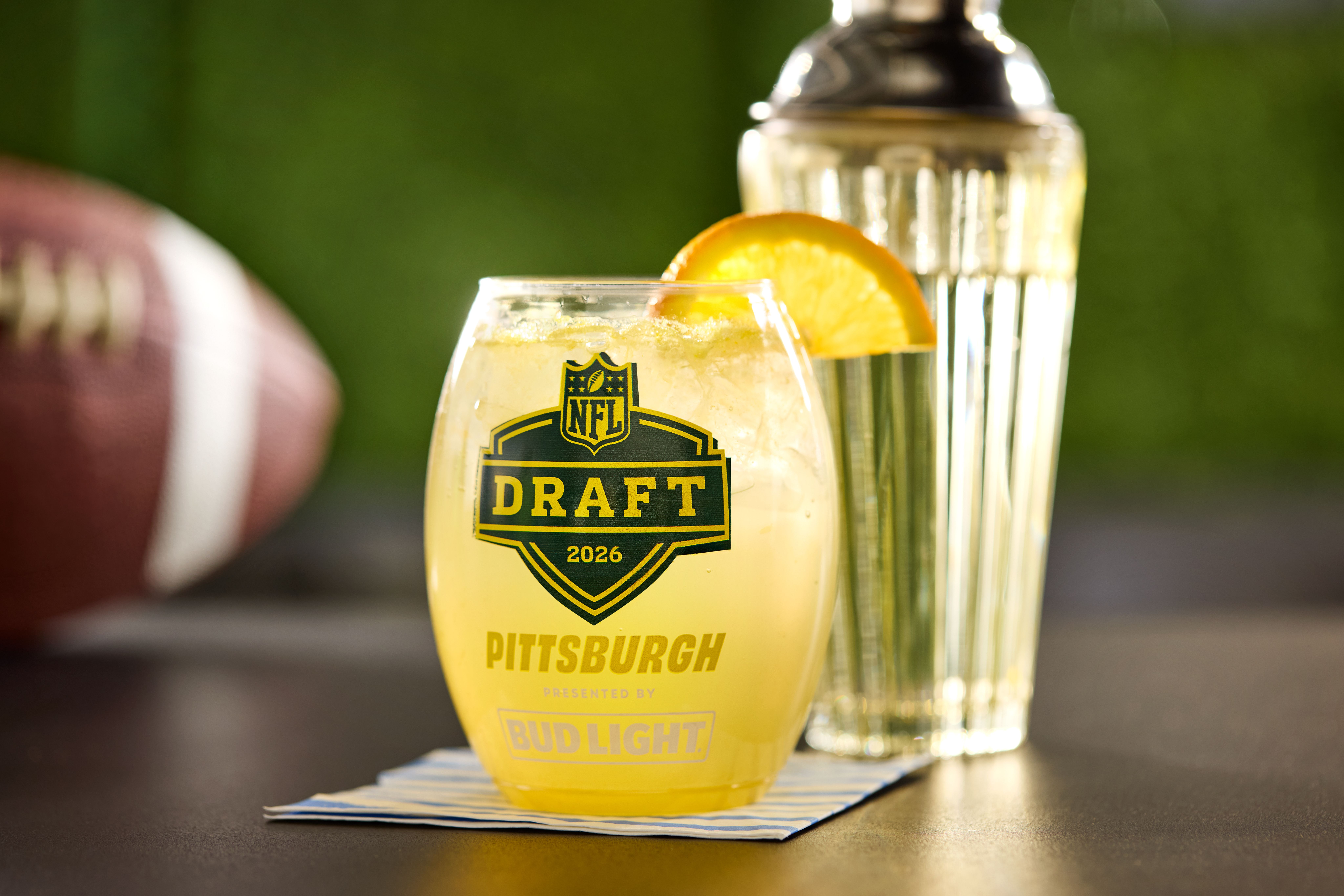 Close-up of a yellow cocktail with an orange wedge on the rim, in a glass labeled "NFL Draft 2026". A tall silver shaker stands behind, on a blue-striped napkin, against a green blurred background.
