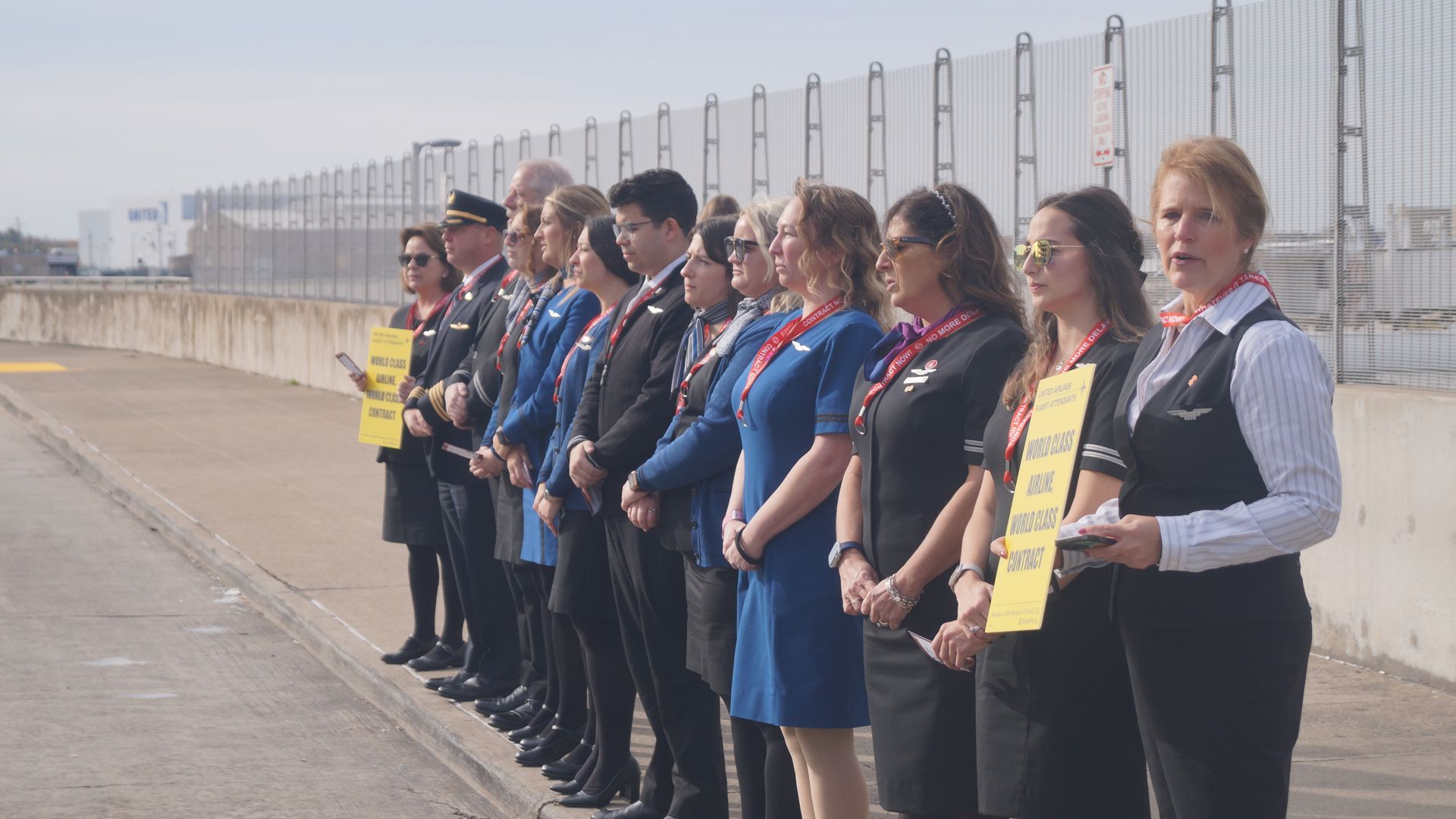 A line of airline flight attendants lined up outside an airport, holding yellow signs. 