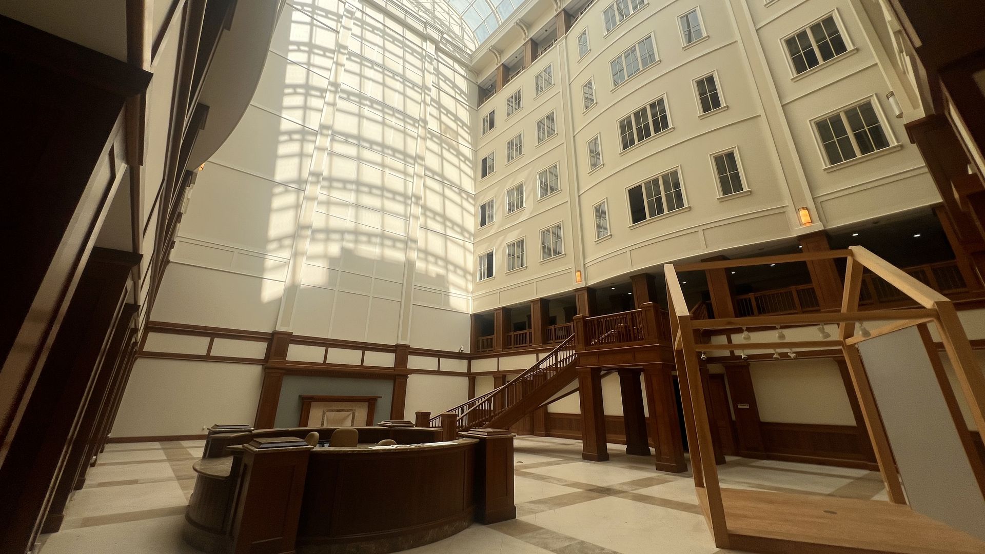 A bright indoor atrium with a tall arched glass skylight, cream walls, and dark wooden railings. Multi-story balconies and stairs surround the space; a circular wooden desk sits in the foreground.
