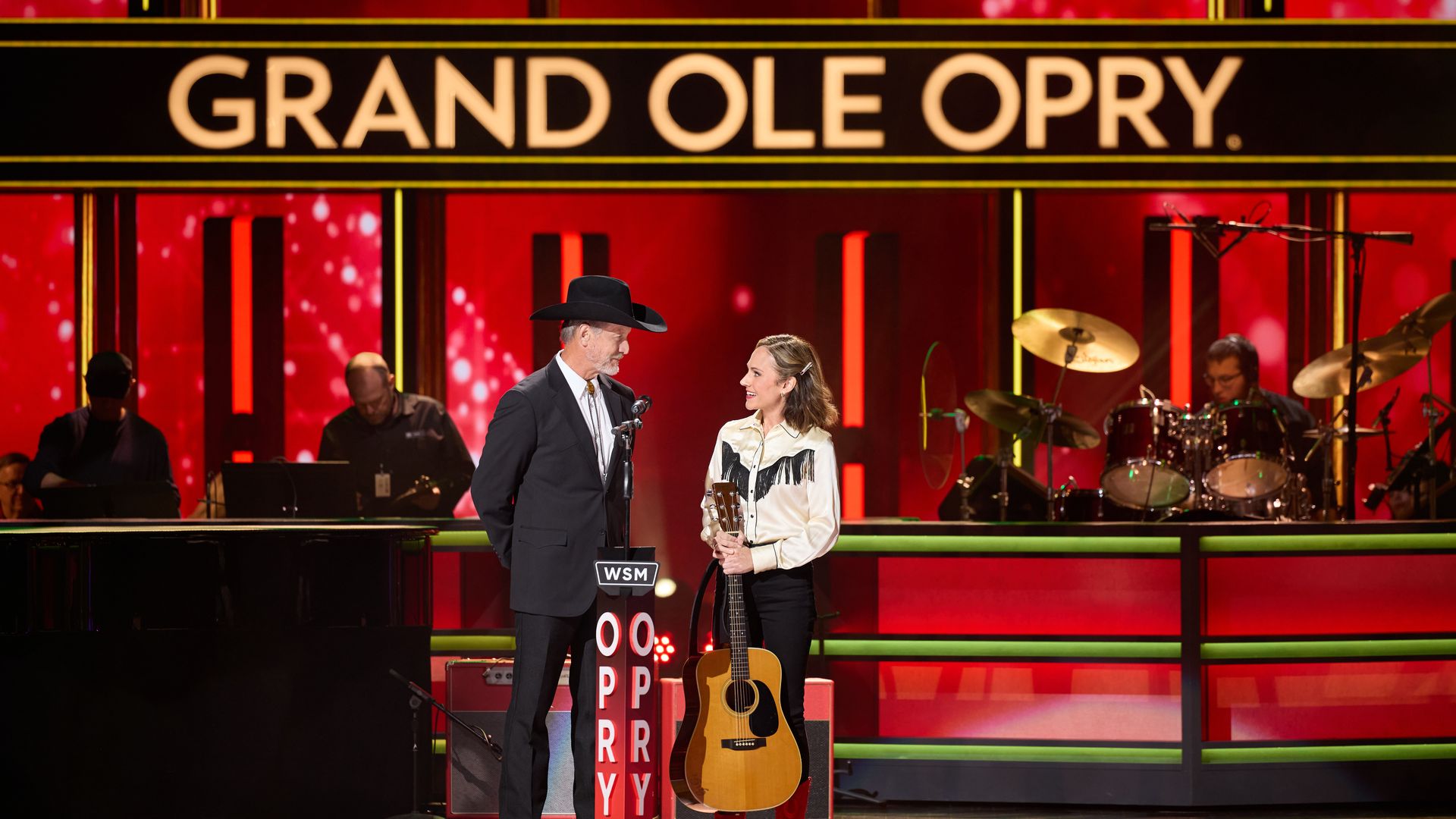 Two people on the Grand Ole Opry stage with a bright red and gold backdrop. A man in a black suit and cowboy hat stands beside a woman holding a guitar, wearing a white shirt with black fringe and red boots.