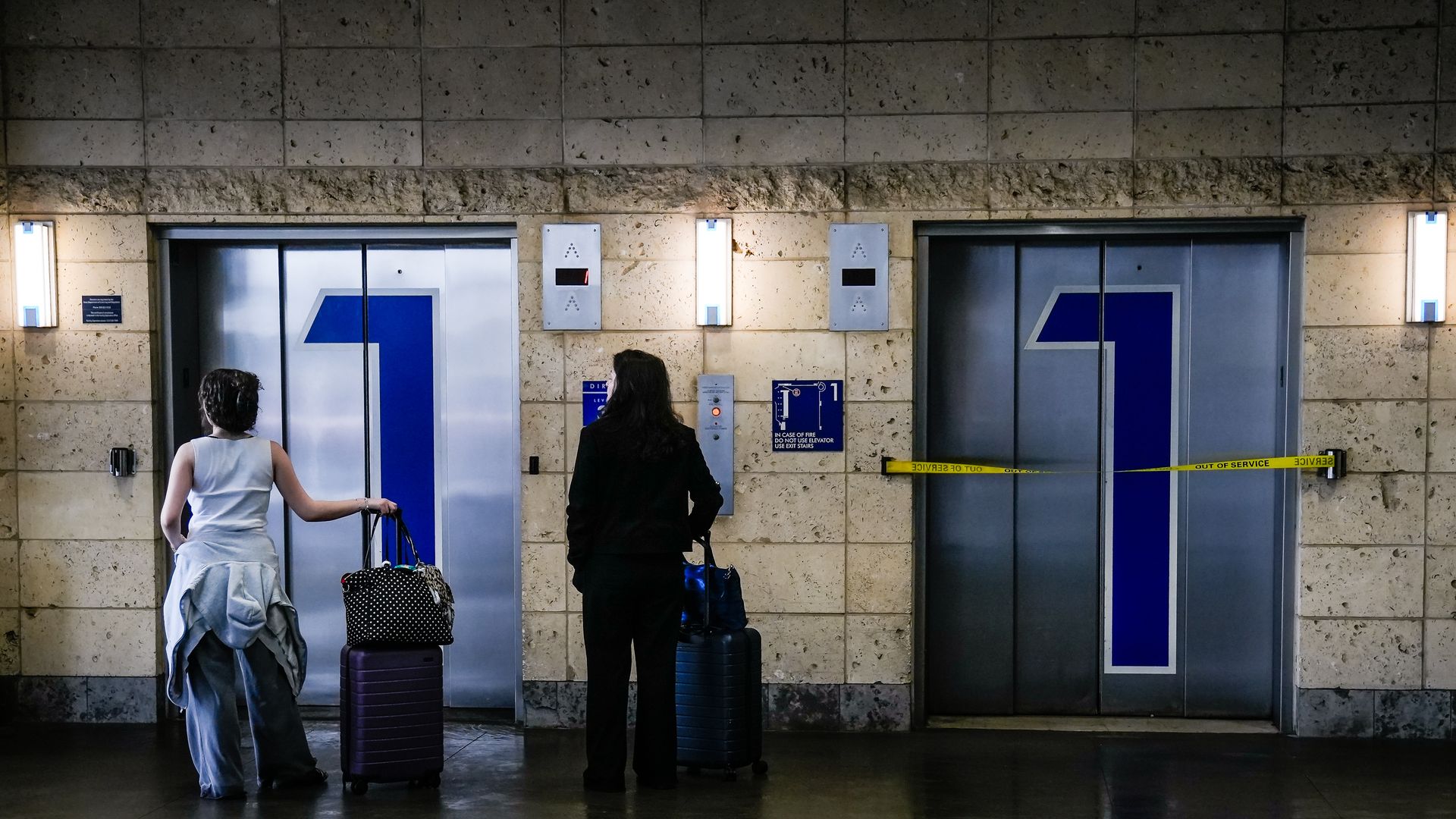 Two travelers with rolling luggage stand before two elevator doors labeled 1 in a station; left, a woman in light blue, right, a person in black with a blue suitcase; yellow tape across the right door.