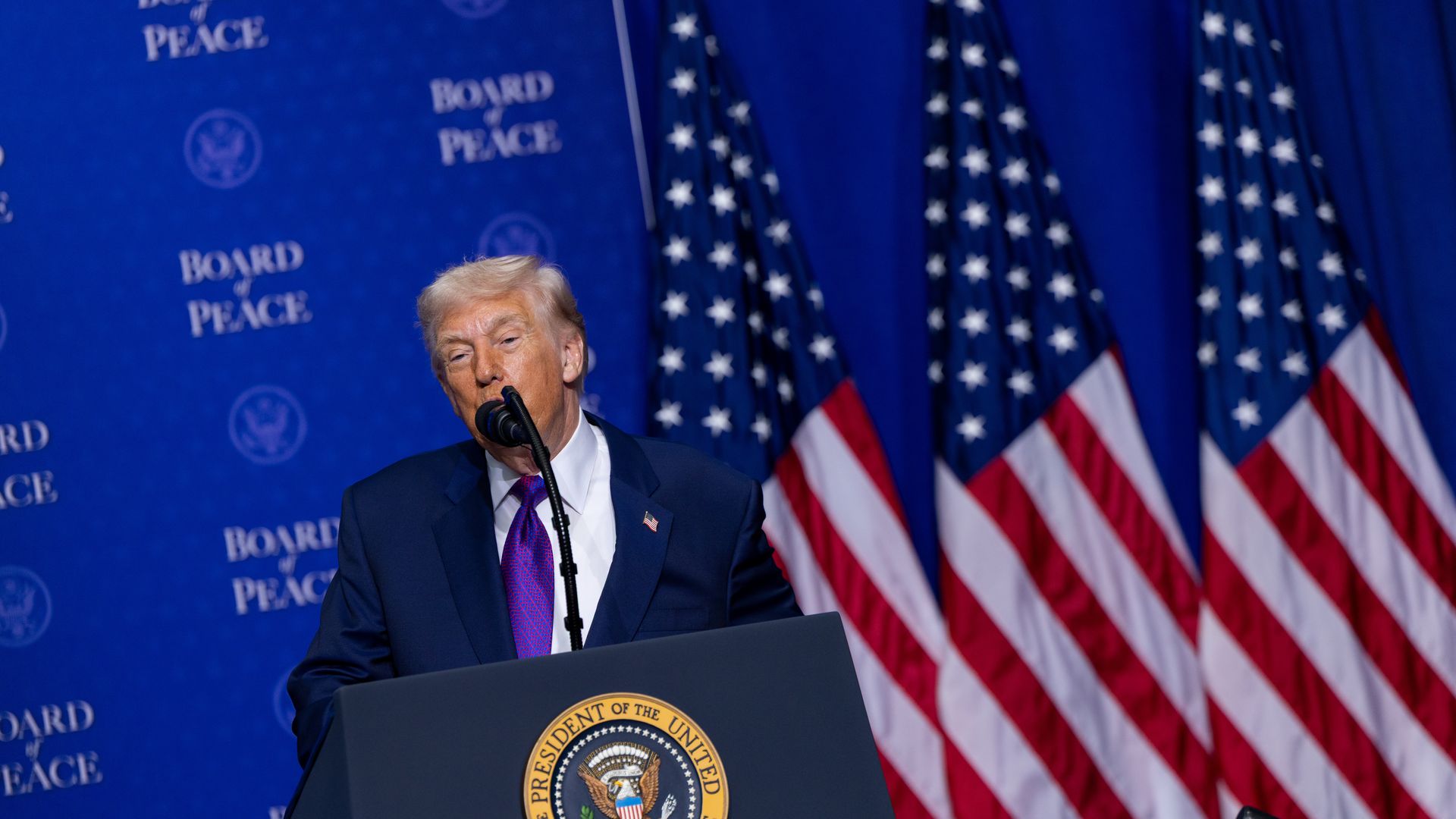 Trump speaks at a lectern with three American flags visible behind him and a backdrop that reads "Board of Peace." 
