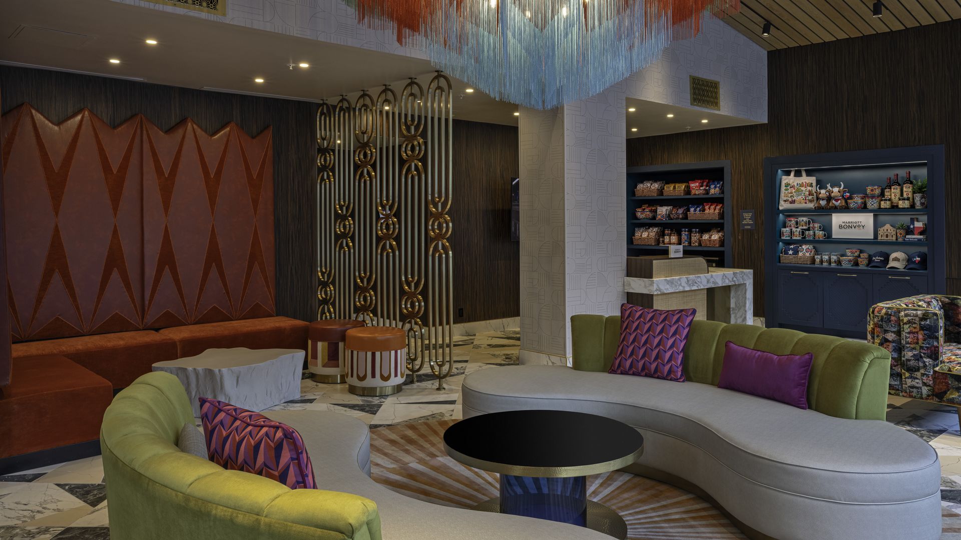 Colorful hotel lobby with curved lime-green and gray sofas, purple cushions, and a round black coffee table. A large multicolor fringe chandelier hangs from the ceiling amid patterned walls and a blue snack shelf.