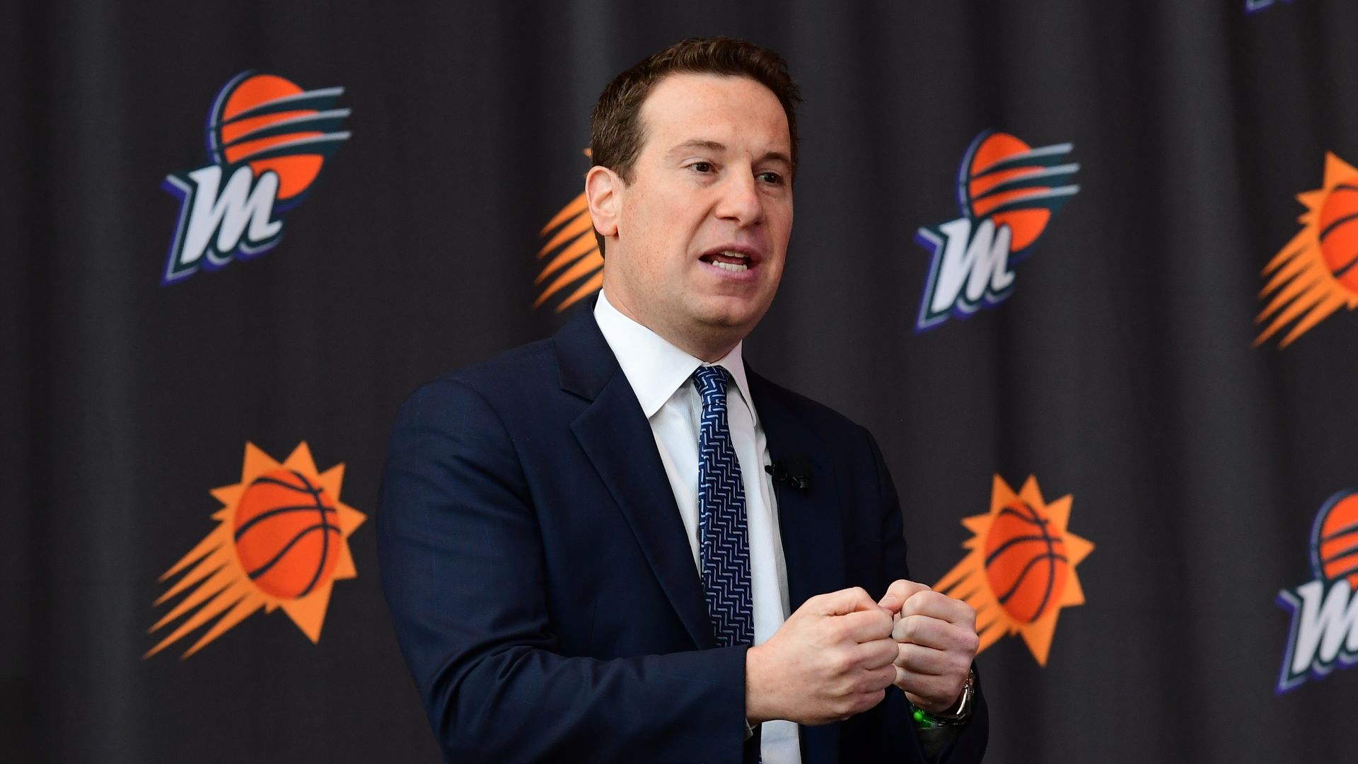 Mat Ishbia gestures with his hands while speaking in front of a backdrop covered in Phoenix Suns and Phoenix Mercury logos.
