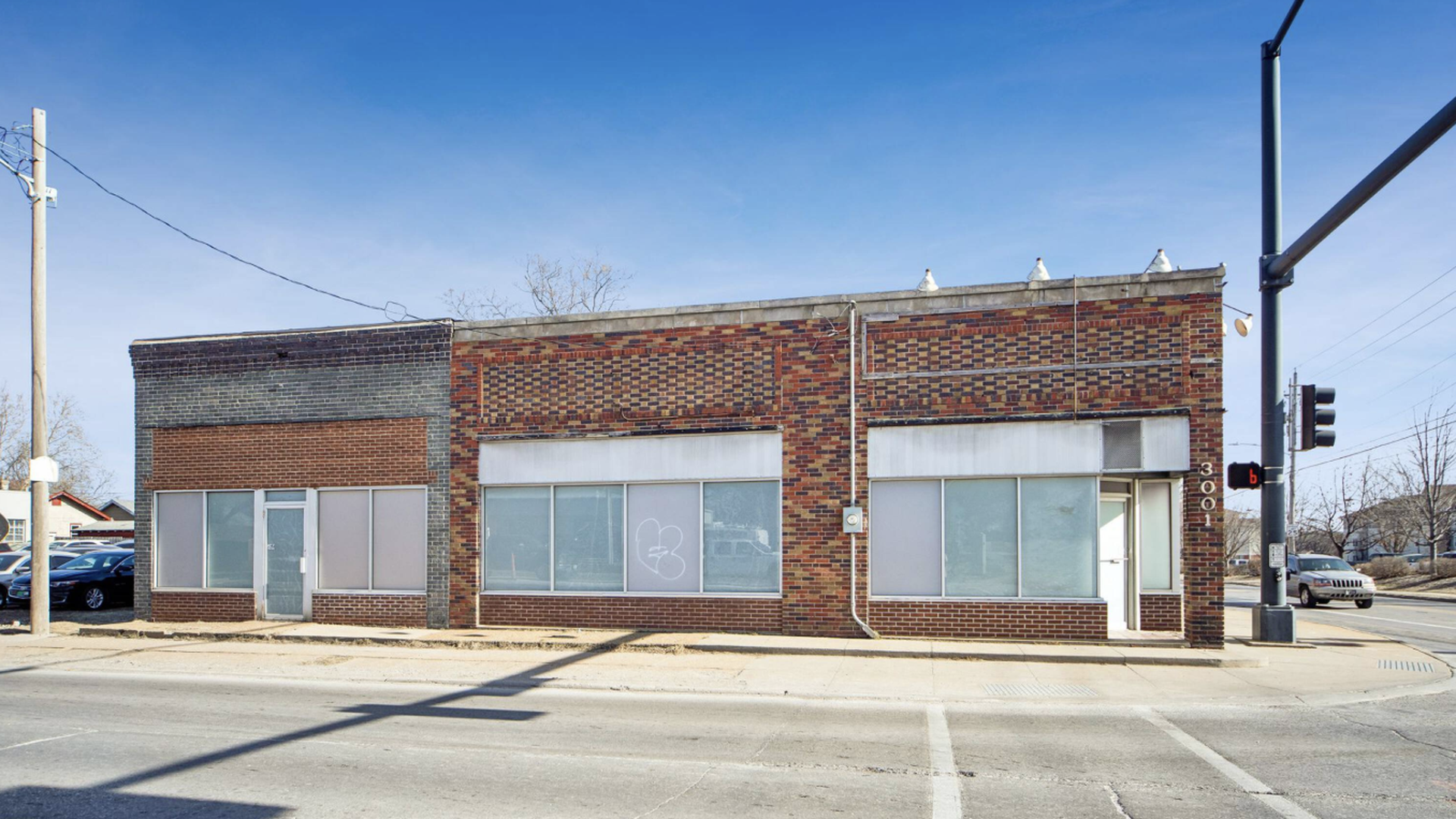 Single-story brick storefront on a street corner under a clear blue sky. Red and gray brick facade with large windows, a white door on the right, and a traffic-light pole casting shadows.
