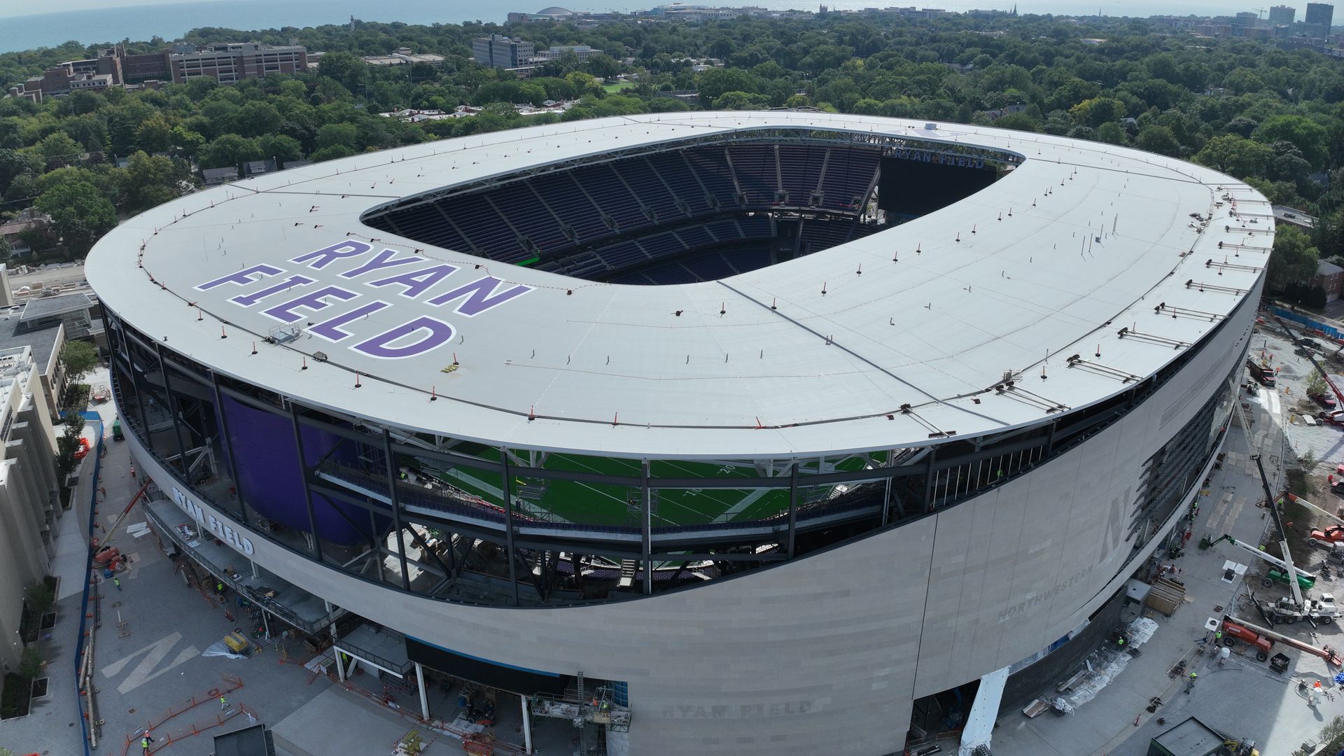 Aerial view of Ryan Field stadium under construction, circular white roof with purple RYAN FIELD text, green trees around, city skyline and a lake on the horizon.