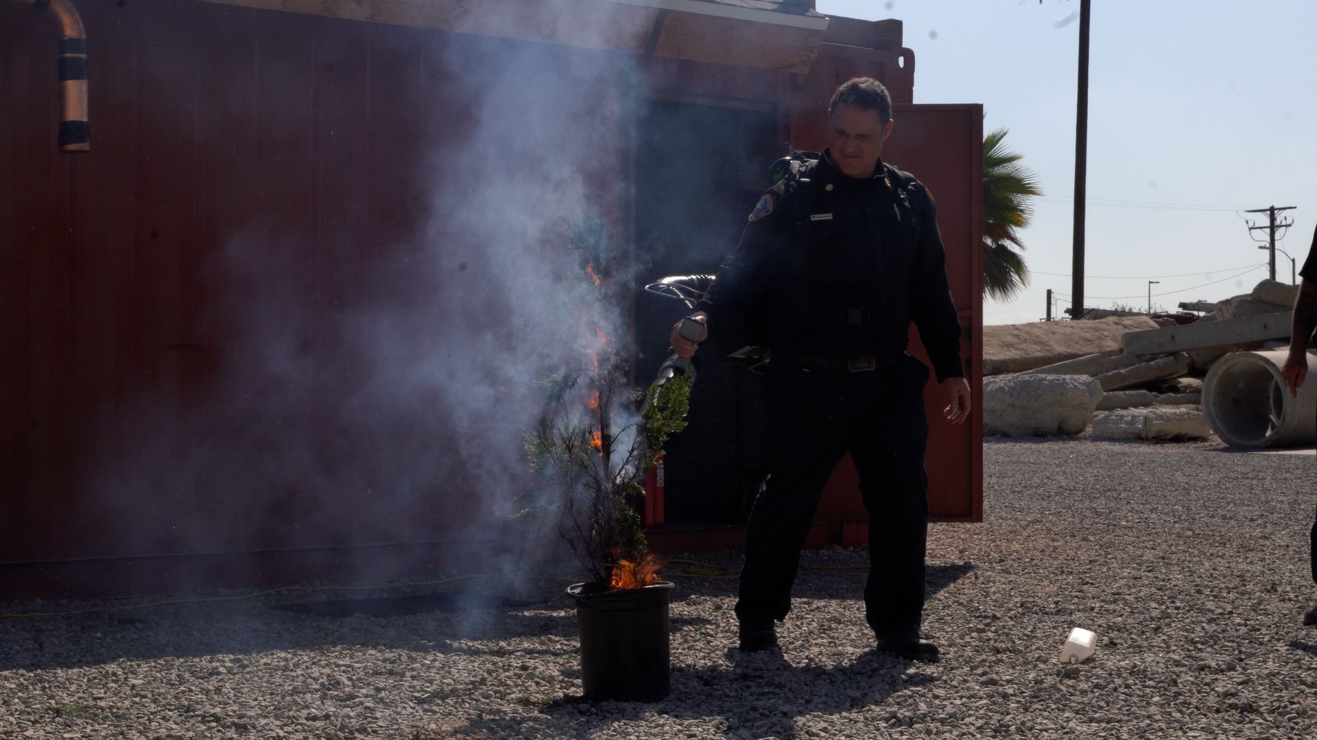A uniformed officer stands on gravel beside a red structure, holding a tool as smoke and small flames rise from a potted plant in a bucket; palm tree and power lines in the background.