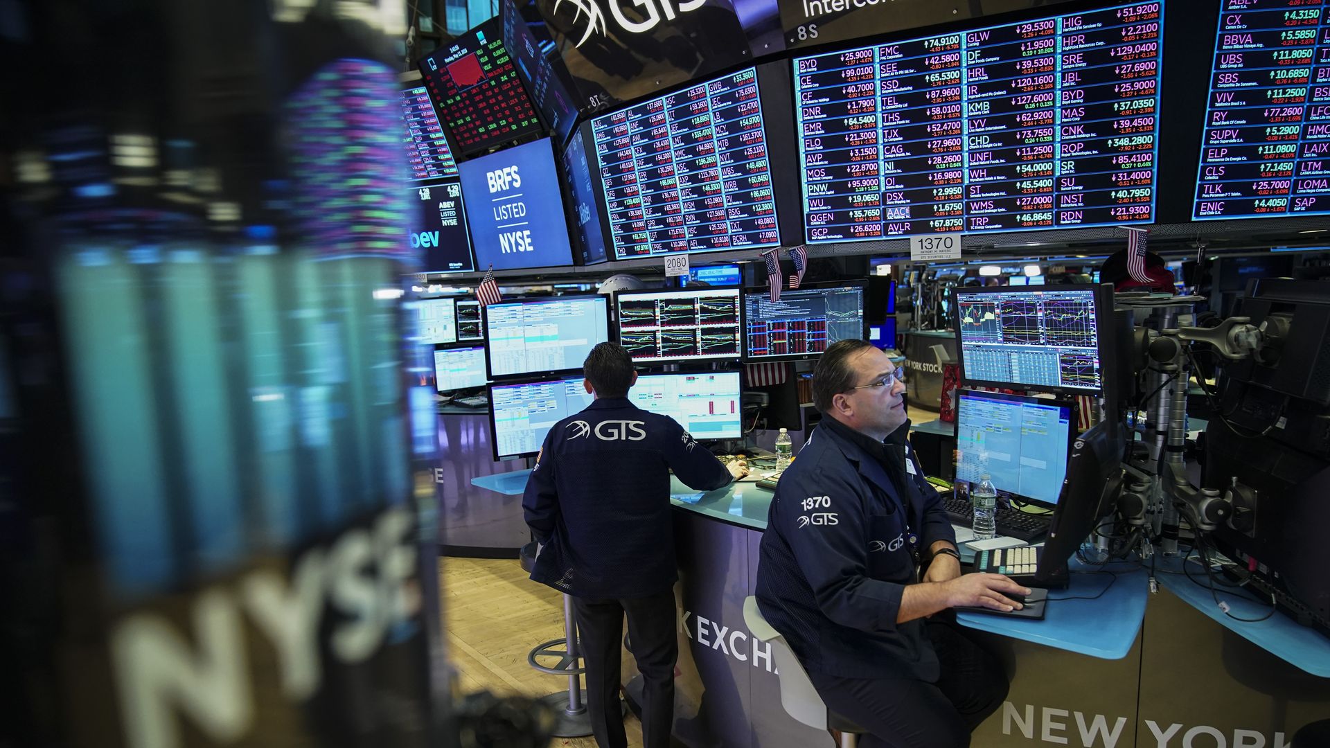 Stock traders sit at desks with screens of company stock tickers overhead.