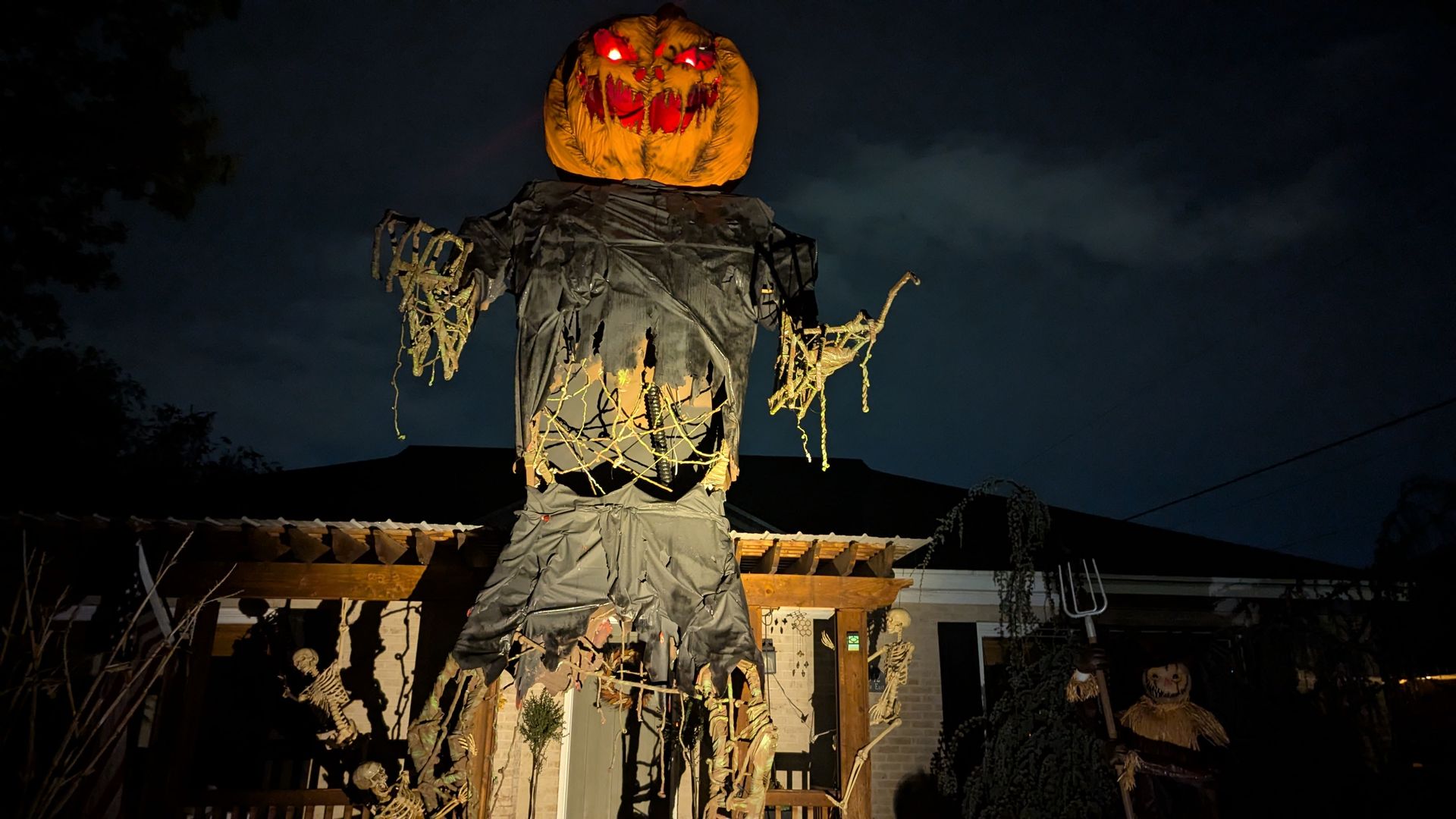 A massive scarecrow with a pumpkin head towers over a house. 