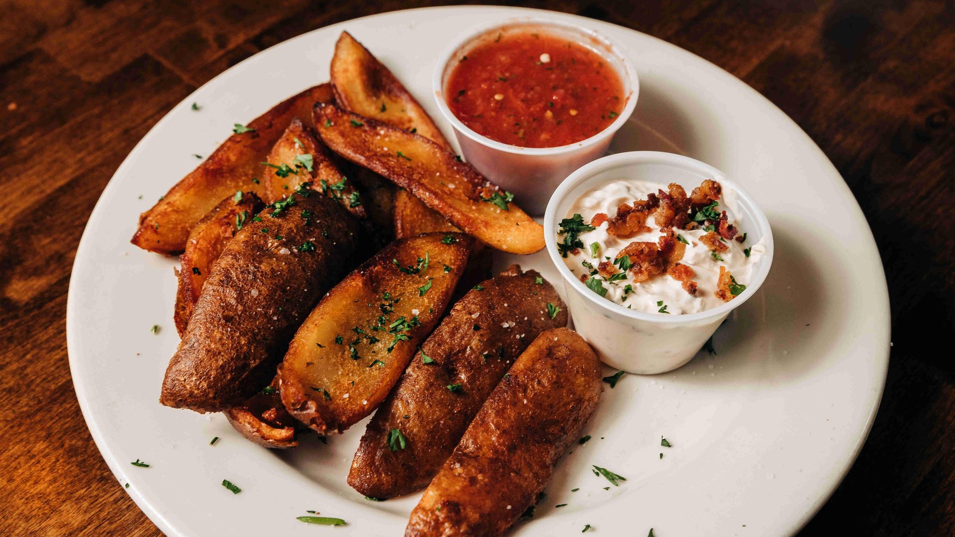 Plate of crispy potato wedges garnished with herbs, served with a cup of red salsa and a cup of creamy white sauce topped with bacon bits and parsley on a wooden table.