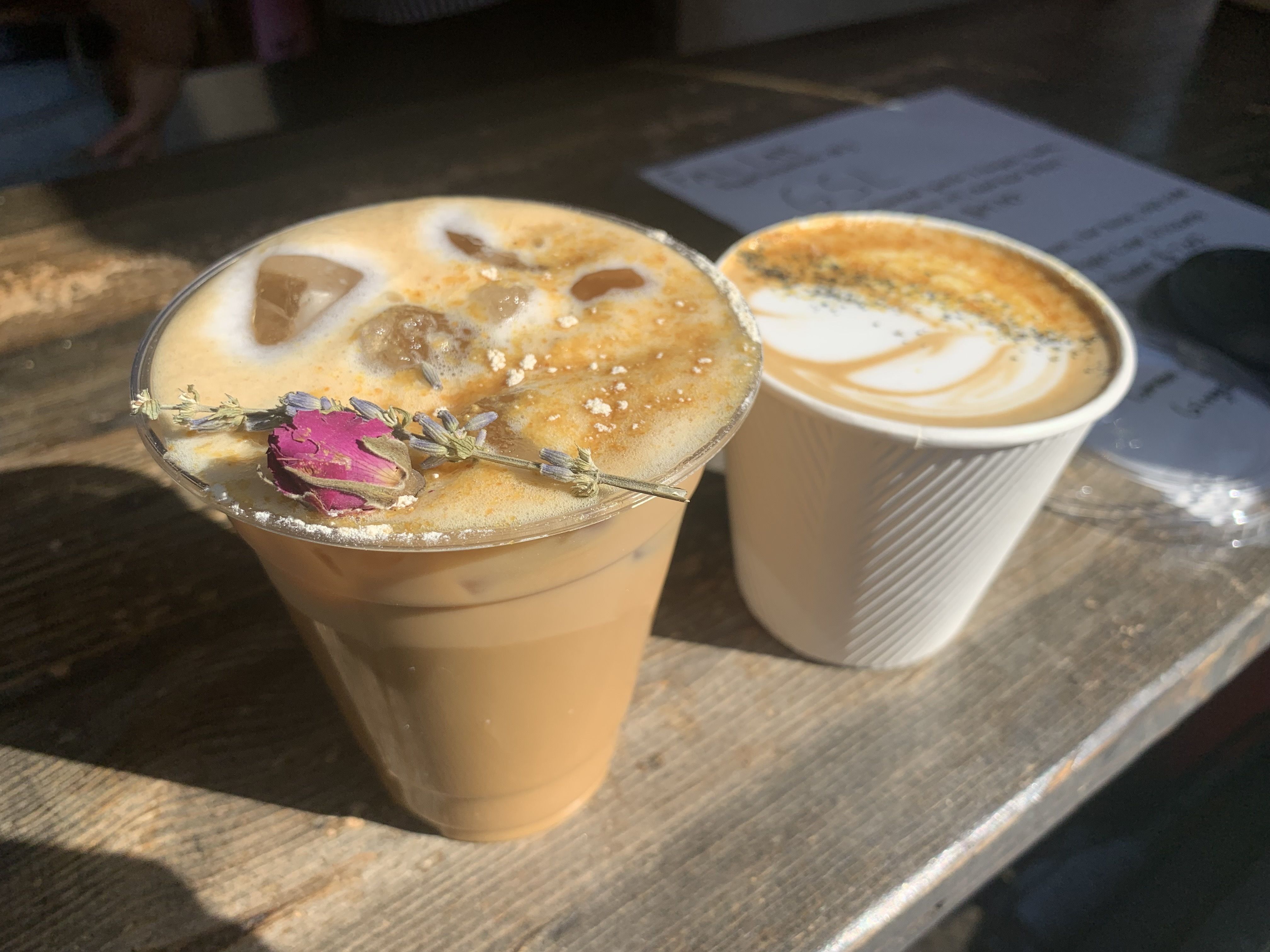 Two coffee drinks on a wooden table: an iced latte with foam and a dried rose garnish, and a hot latte in a white cup with frothy art and sprinkled spices.