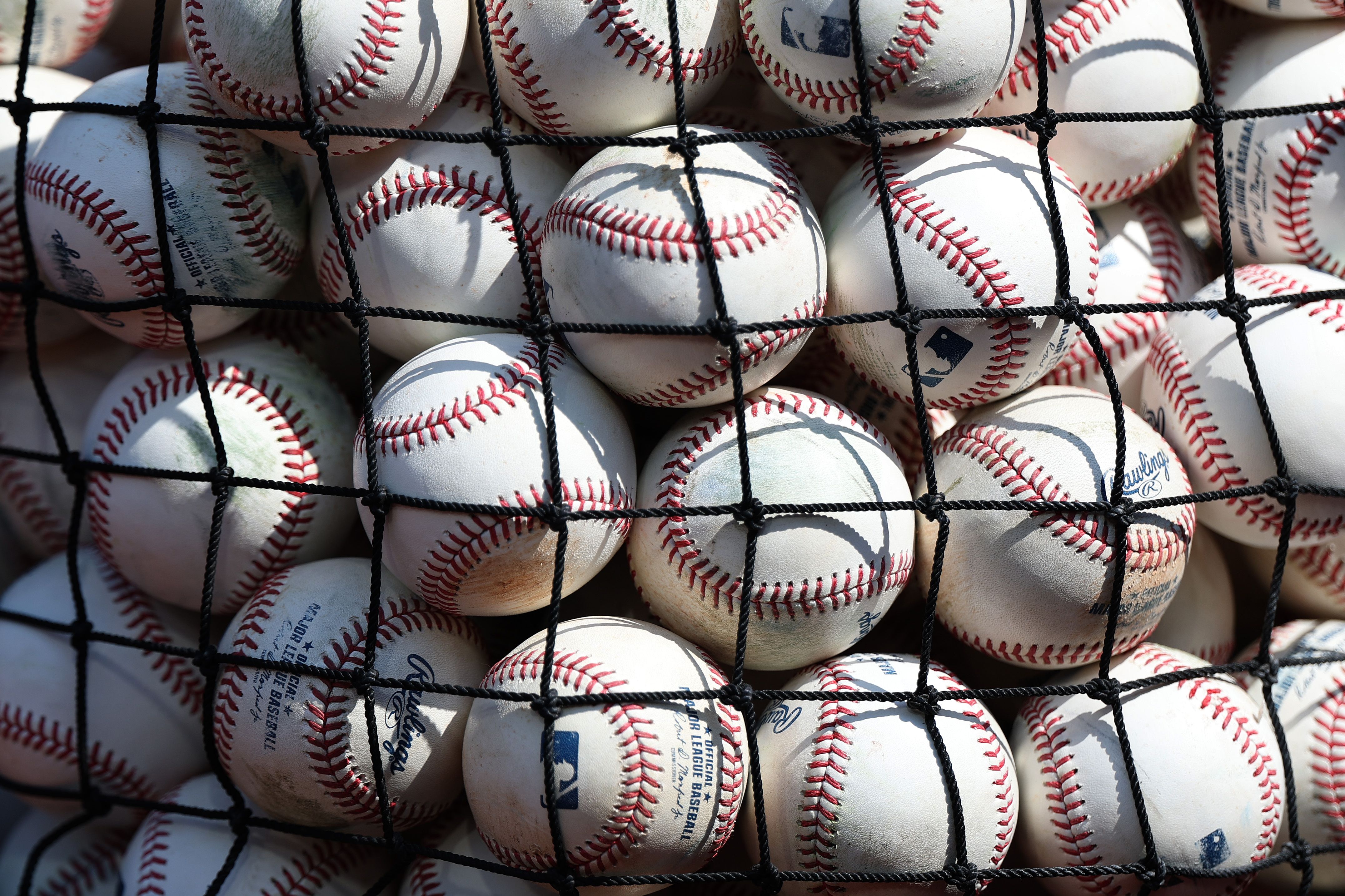 Close-up of white baseballs with red stitching piled behind a black net, some showing the MLB logo and Rawlings branding, with shadows across the raised seams.