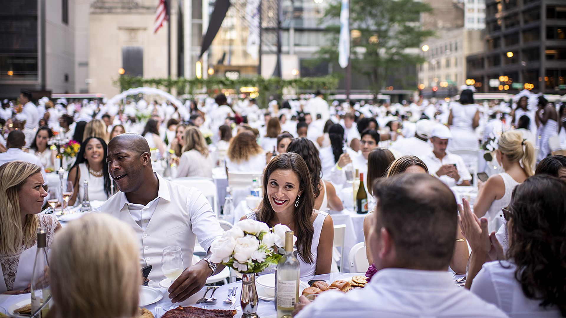 Crowd of people dressed in white sitting together at a dinner table with white flowers on table.