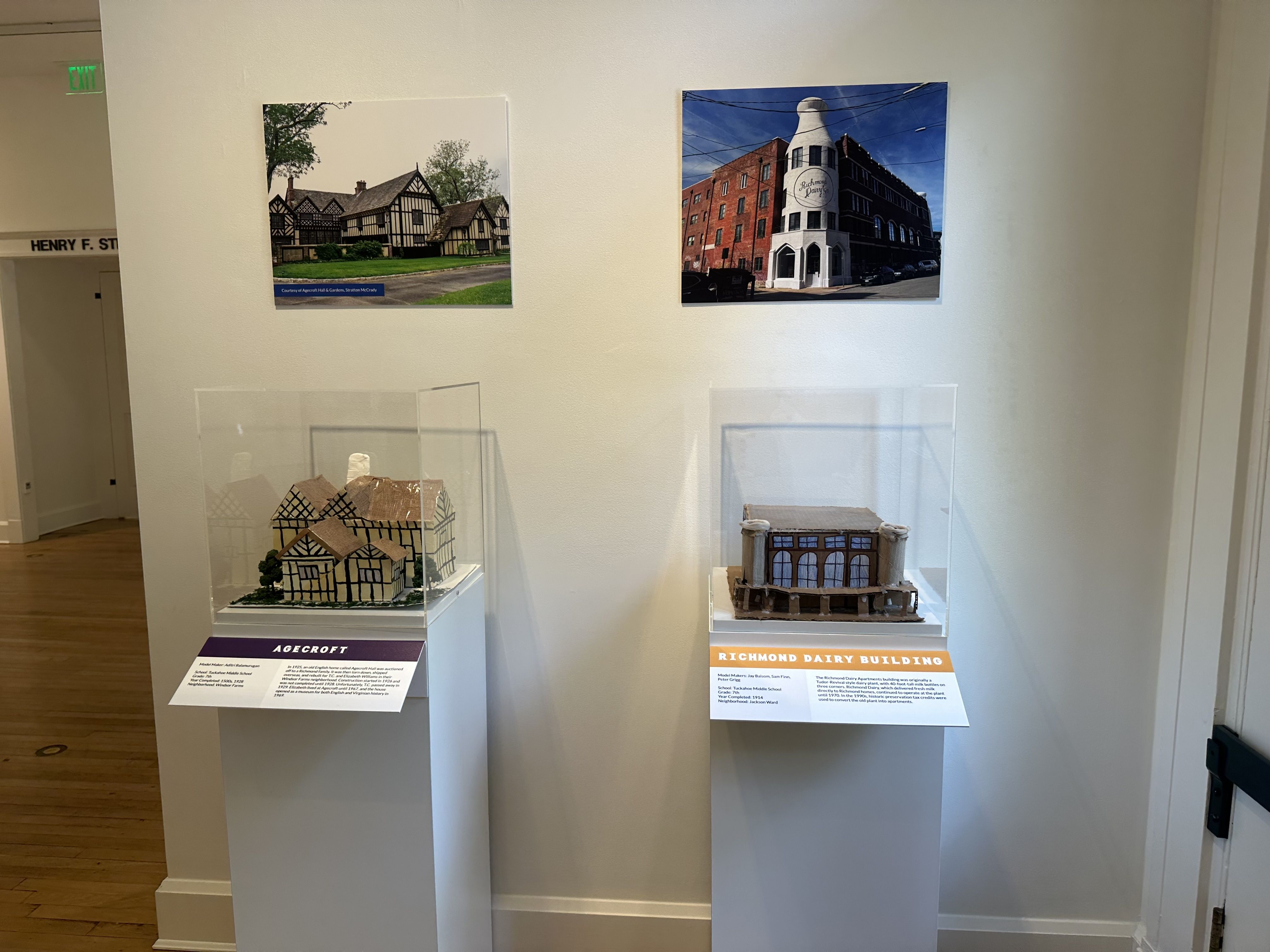 Two wall photos in a gallery: left shows Tudor-style houses with a green lawn; right features a tall white corner tower on a red brick building labeled Richmond Dairy Building under a blue sky. Scale models sit in clear display cases below.