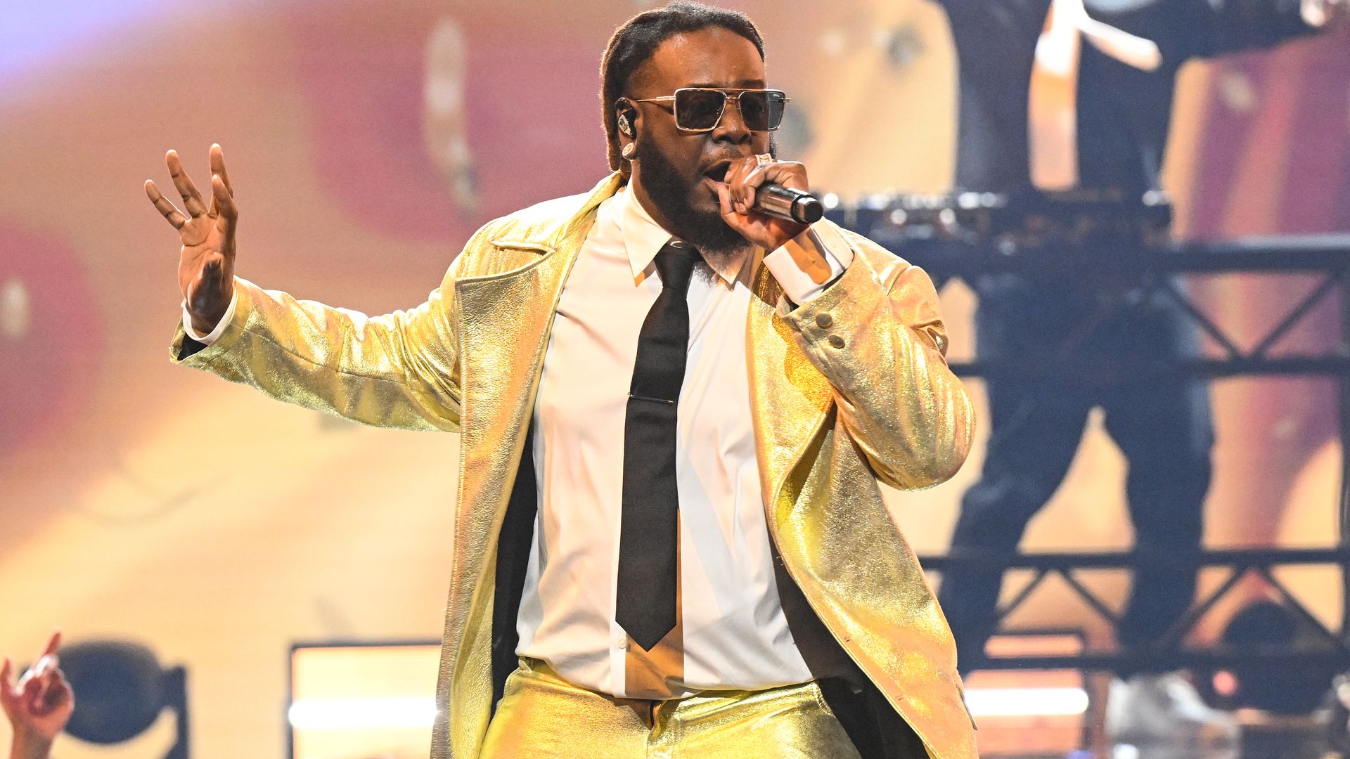 T-Pain sings into a microphone on stage wearing a suit and tie. 
