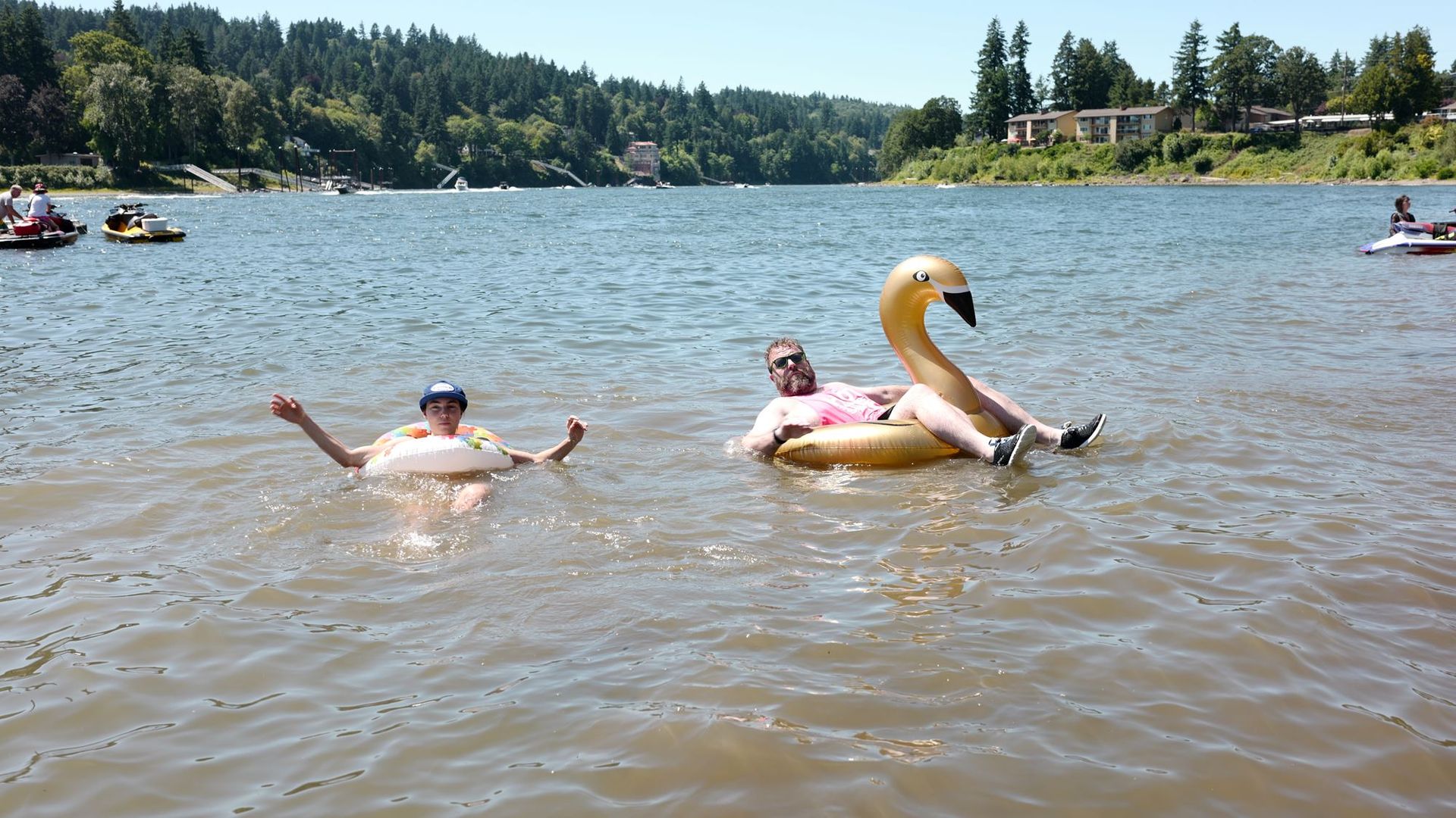 Two people floating on a lake in inflatable tubes—one on a white tube, the other on a gold swan tube. Green tree-covered shoreline and a few buildings visible in the background.