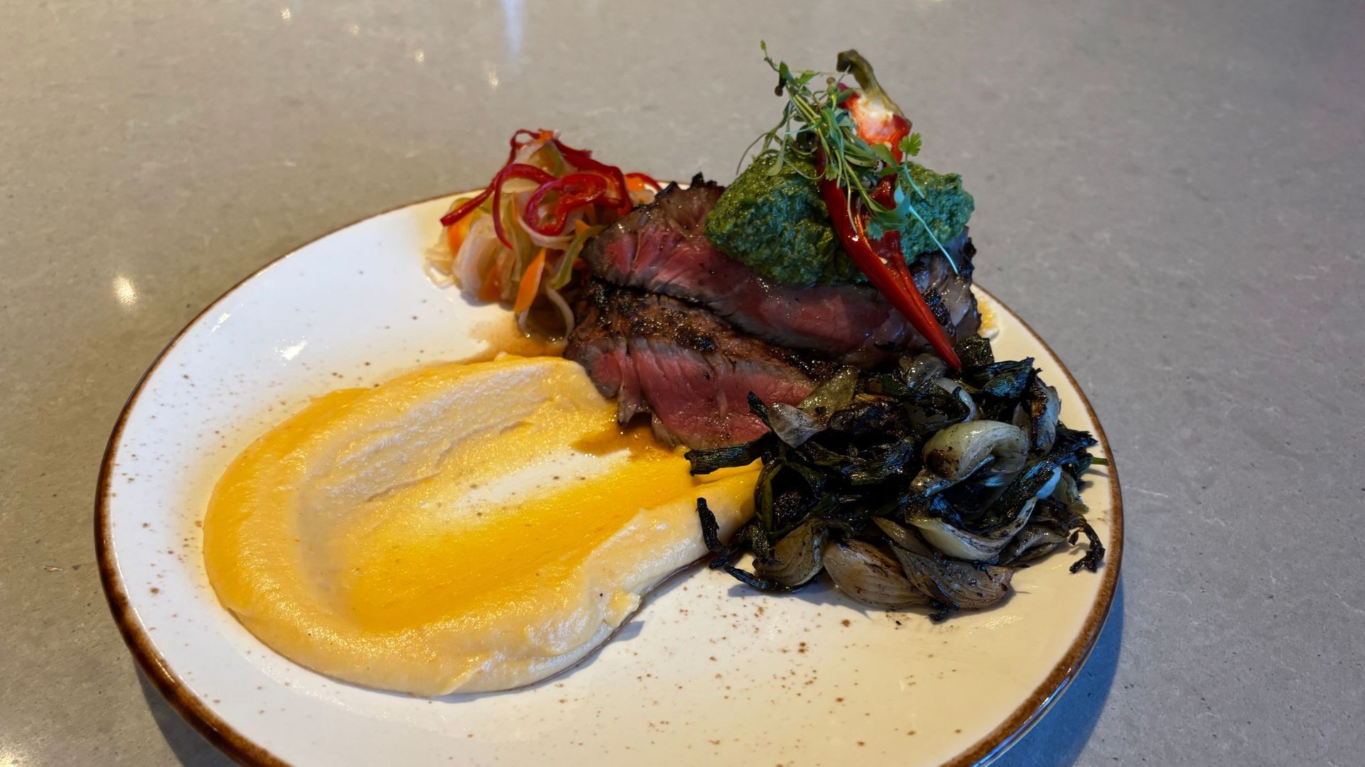 Plate with slices of medium-rare beef topped by green herb pesto and microgreens, beside creamy yellow mashed potatoes and sautéed onions and peppers on a white plate with a brown rim.