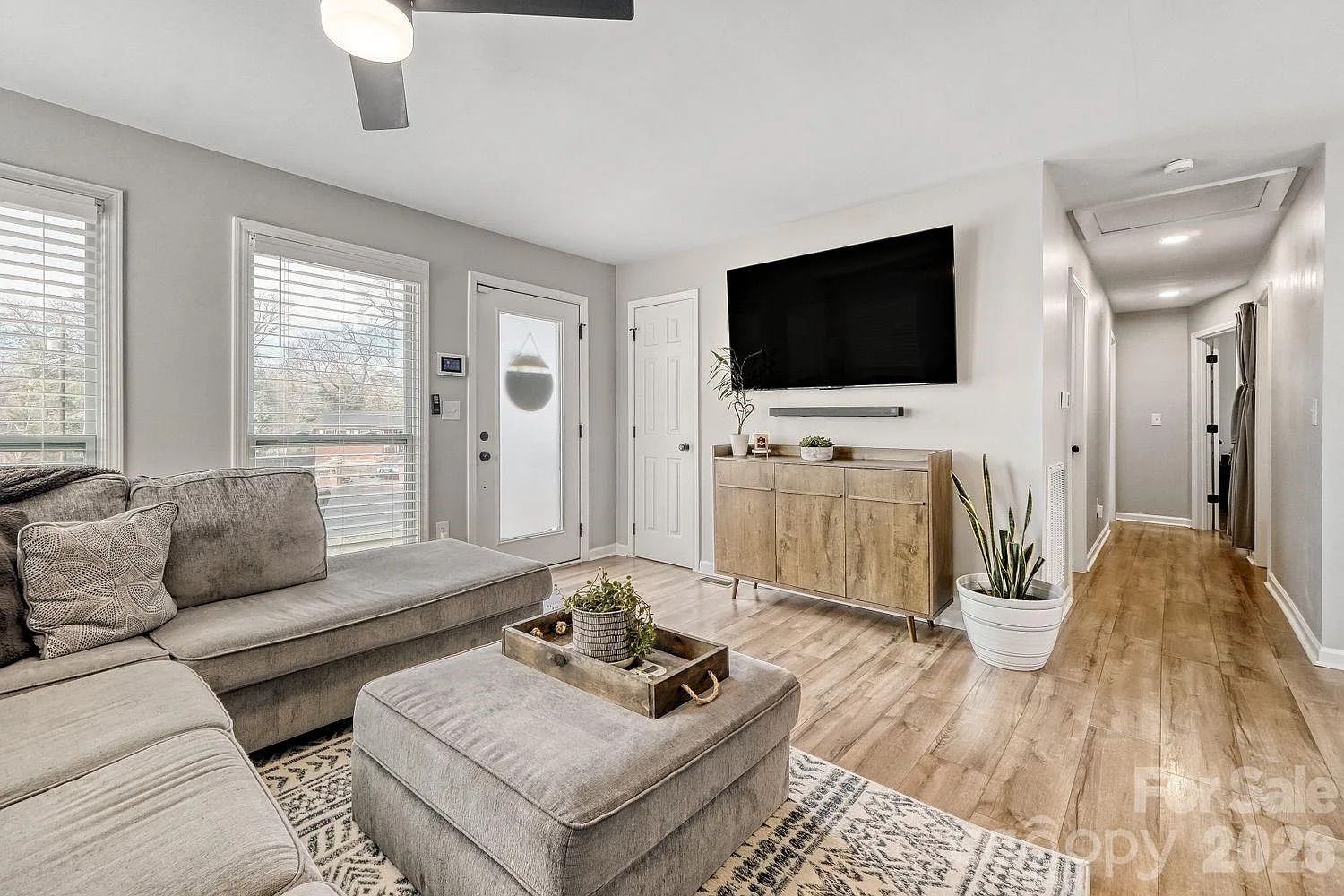 Modern living room with gray sectional sofa, matching ottoman, wooden media console, large wall-mounted TV, light wood flooring, patterned rug, potted plants, and natural light from windows.
