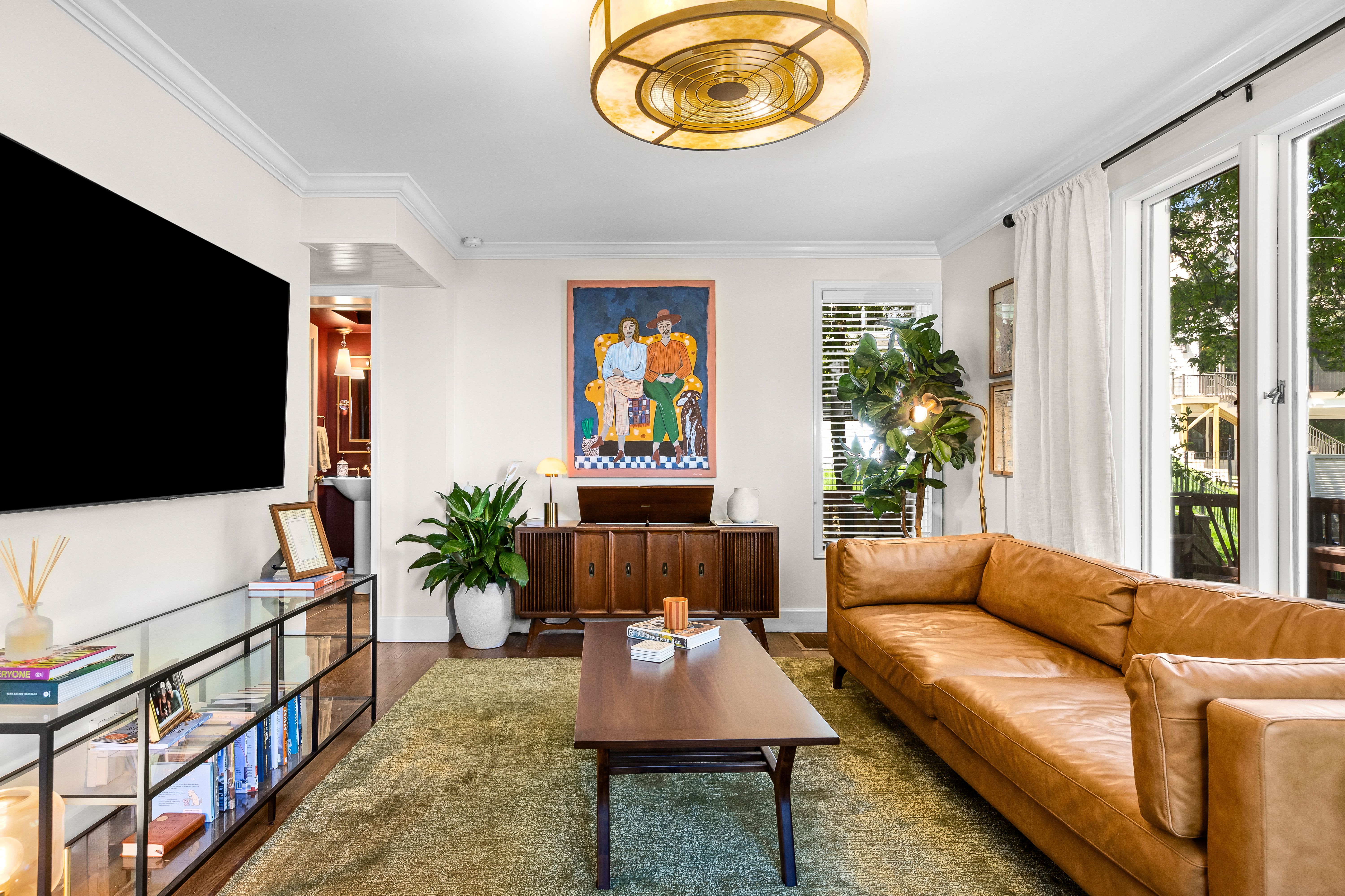Bright living room with a large black flat-screen TV on the left, a wooden console with a colorful painting, green plants, a glass coffee table, a tan leather sofa, and wide windows with curtains.