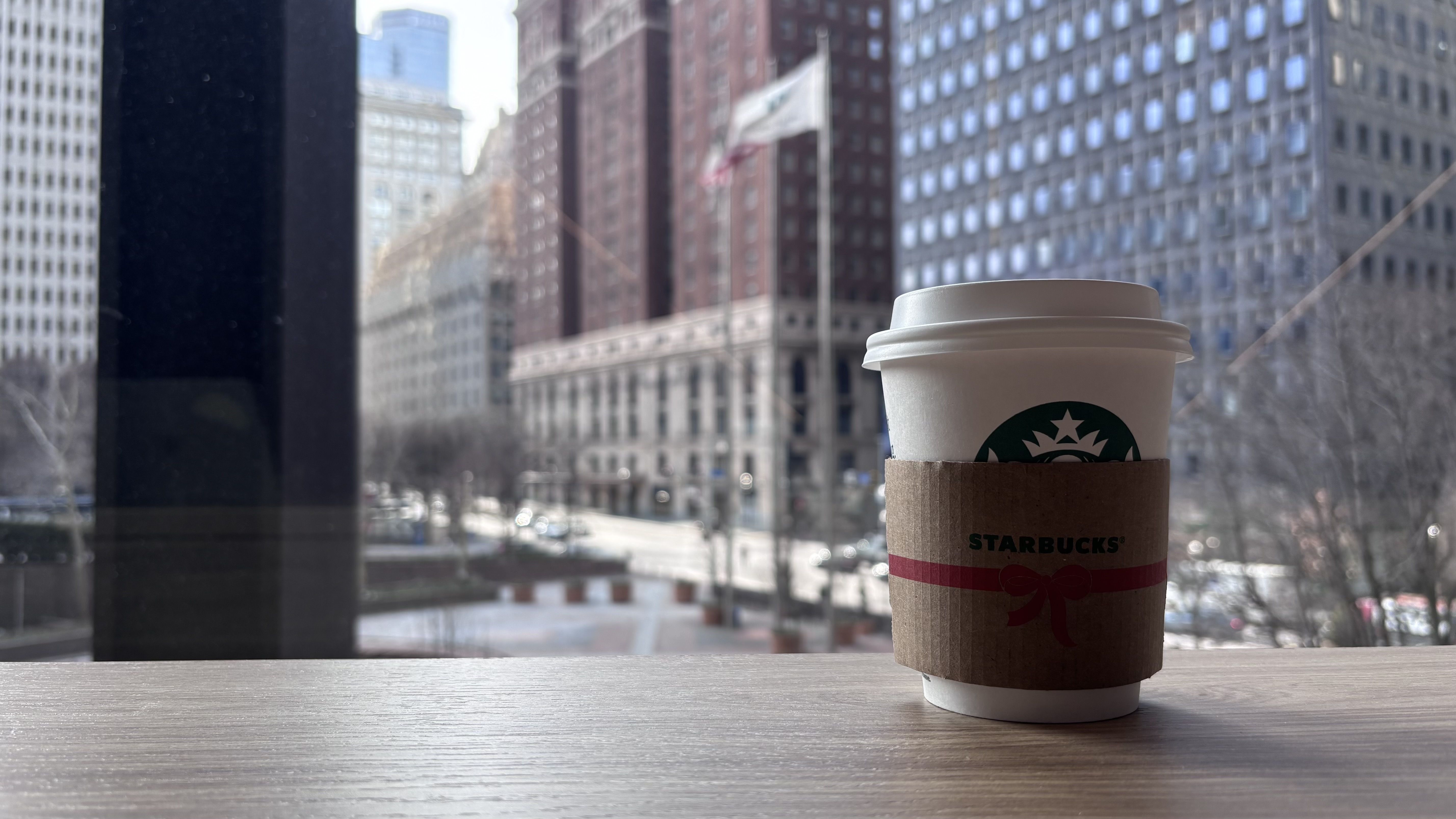 Starbucks coffee cup with brown sleeve and white lid on wooden table, city buildings and flag visible through large window in background during daytime.