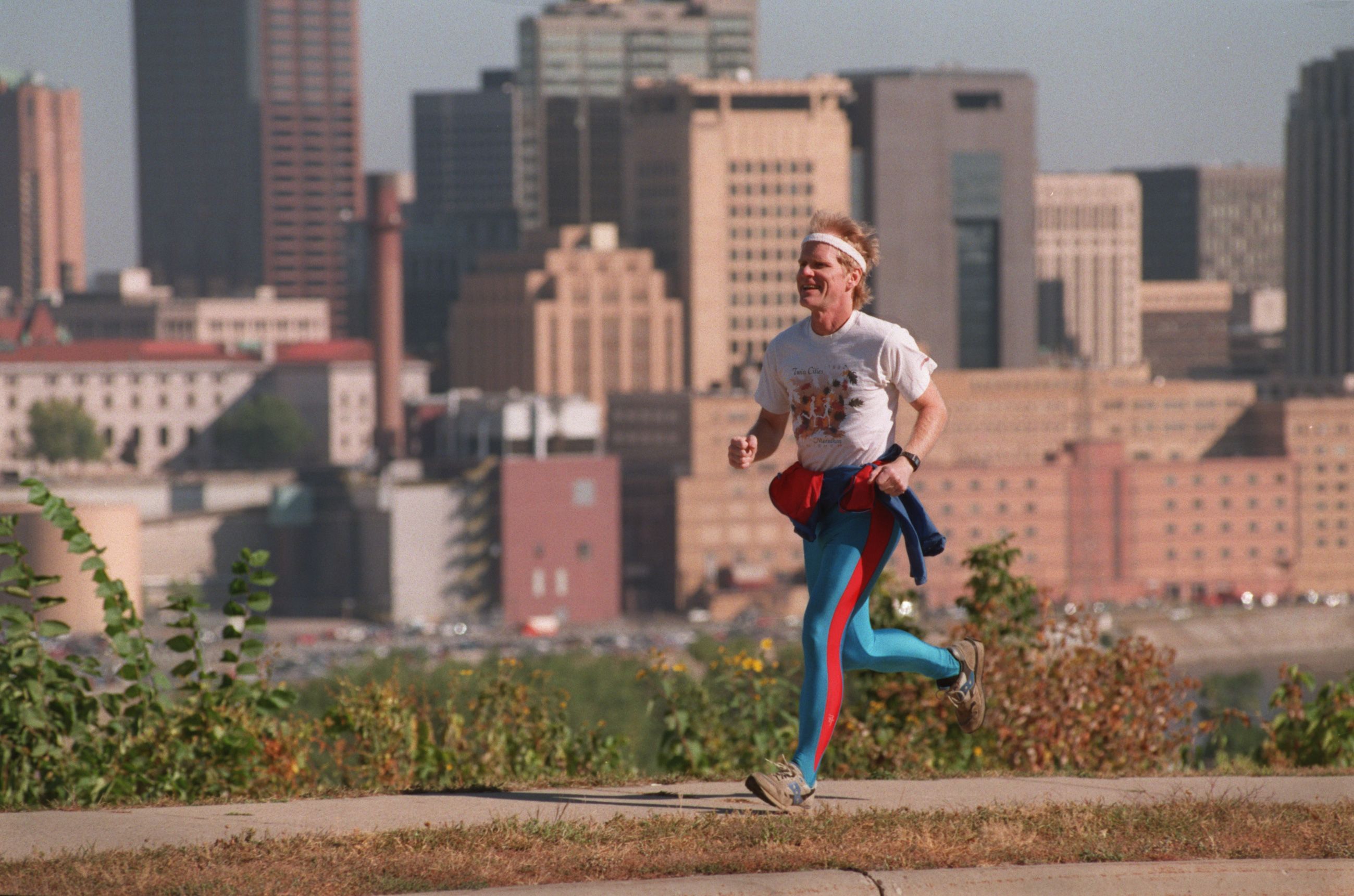 Man running along a city skyline. He wears a white headband and white graphic tee, with bright blue pants and a red jacket tied around his waist, expression joyful as he moves.