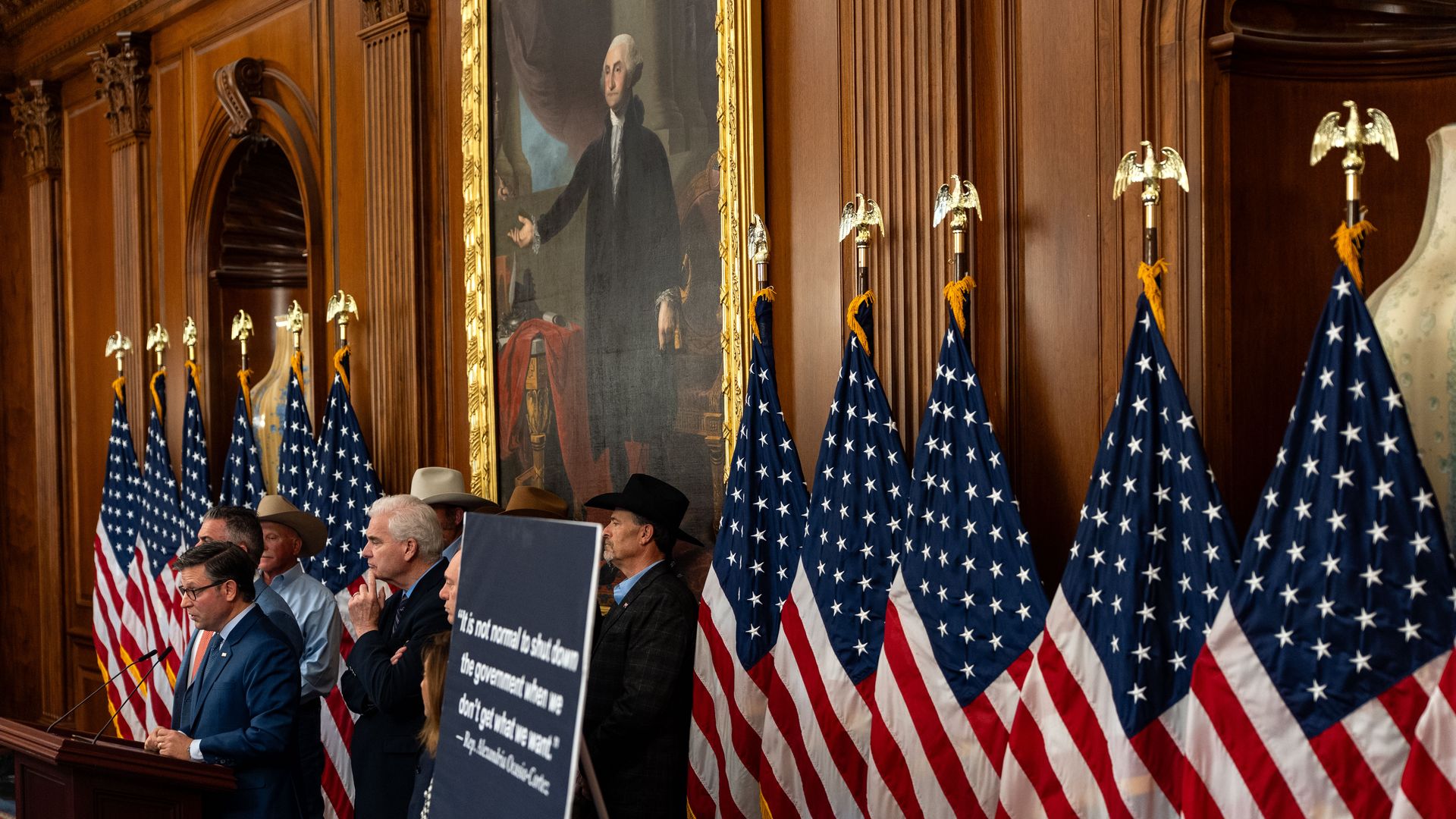 House Speaker Mike Johnson (R-LA) answers questions from the press at the U.S. Capitol, standing in front of many American flags and a sign with a quote from Rep. Alexandria Ocasio-Cortez.
