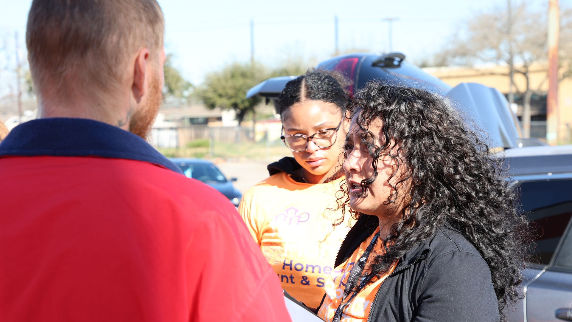 Two women in orange shirts talking to a man in a red jacket outdoors near parked cars, one woman with curly hair holds a folder, sunny day with trees and buildings in background.