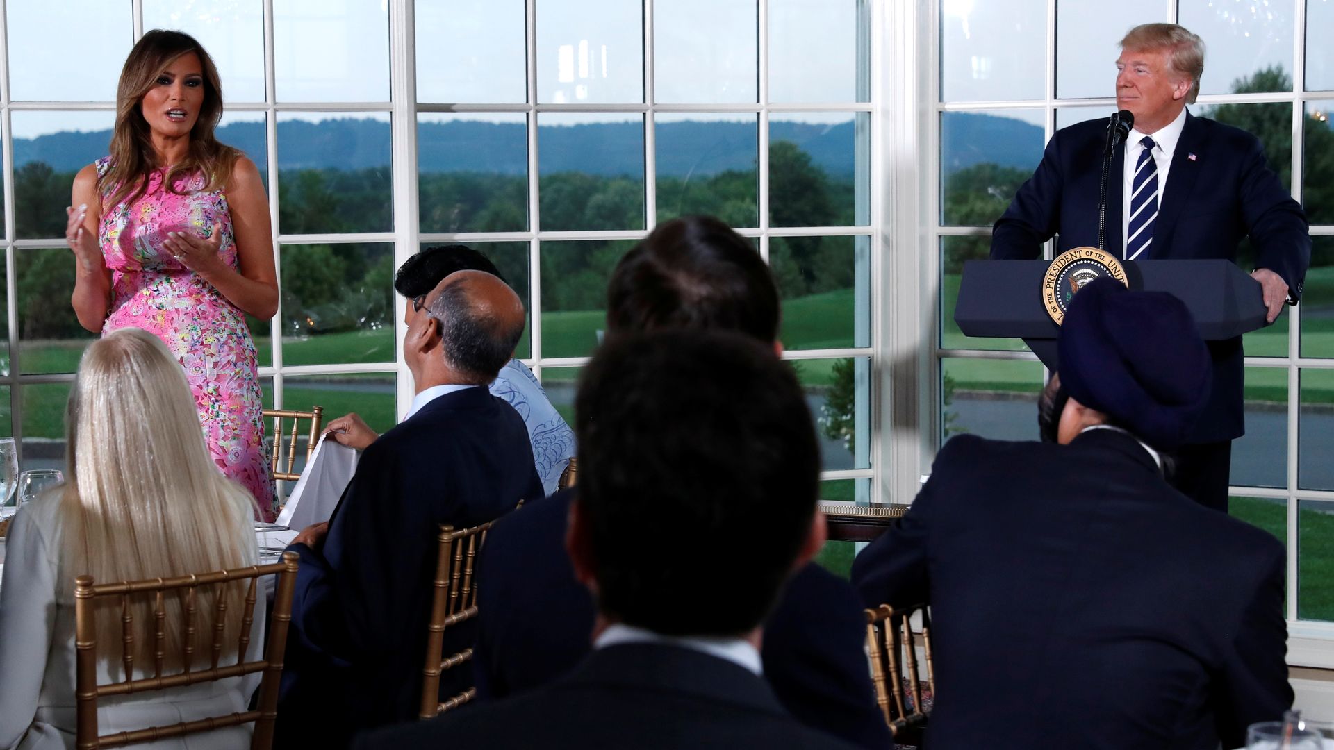Donald Trump and Melania Trump are pictured here speaking in front of seated guests at a dinner, with several windows facing a golf course behind them.