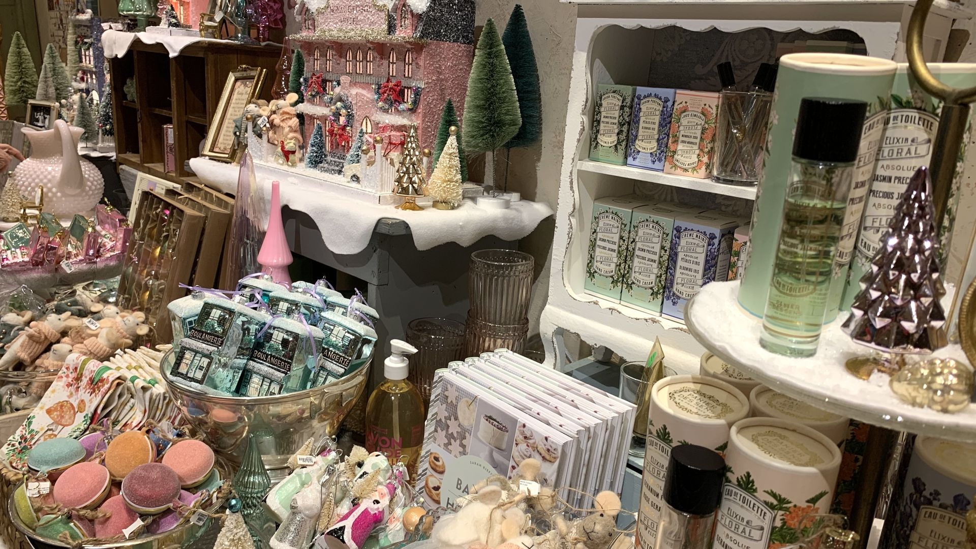 Decorative display with pastel-colored Christmas ornaments, small felt mice, pink and green trees, boxed toiletries, and a glittery miniature village on wooden shelves.
