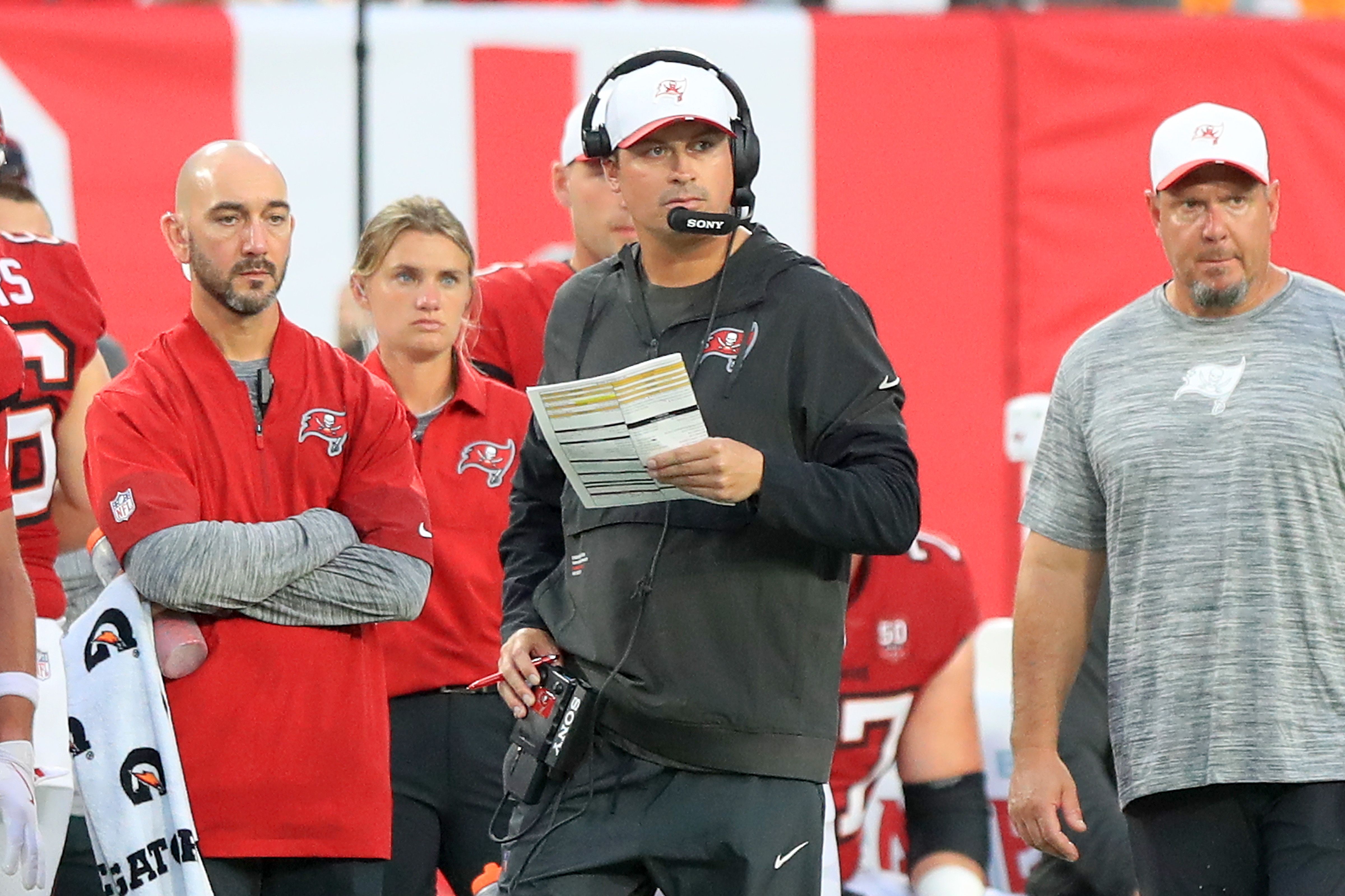 Tampa Bay Buccaneers coaching staff on sidelines in red and gray gear during a football game, one holding a play sheet and wearing a headset with a red and white cap.