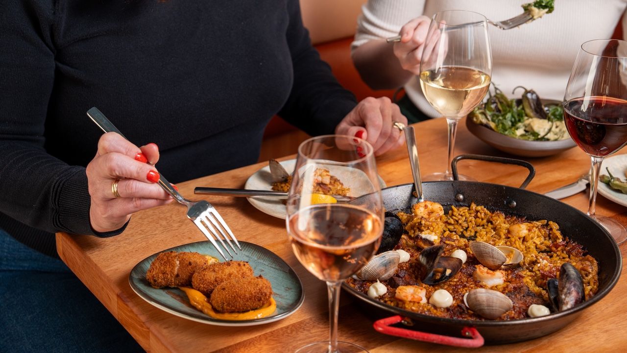 Two people dine at a wooden table: a large pan of paella with shrimp, clams, and mussels, a plate of croquettes with orange sauce, and wine glasses nearby.