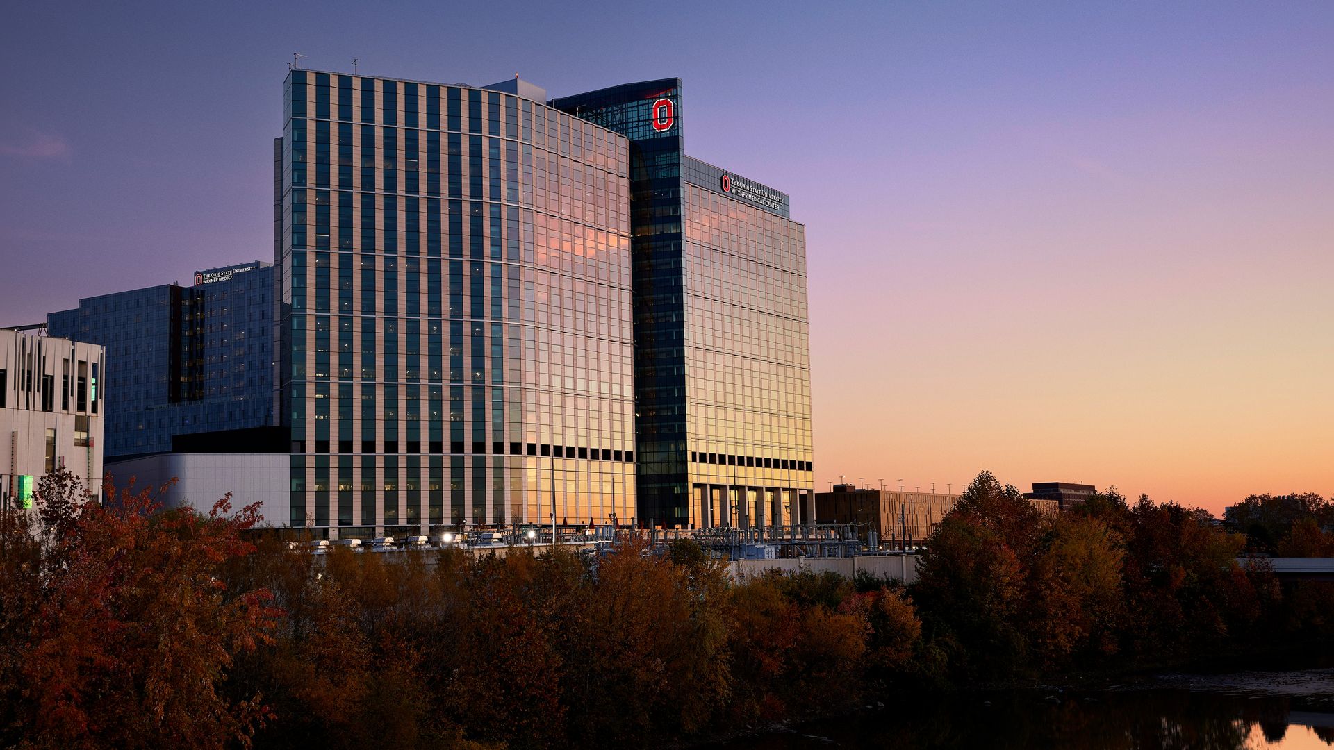 Modern glass buildings of The Ohio State University Wexner Medical Center reflecting sunset hues, with autumn trees and calm water in the foreground under a clear purple-orange sky.