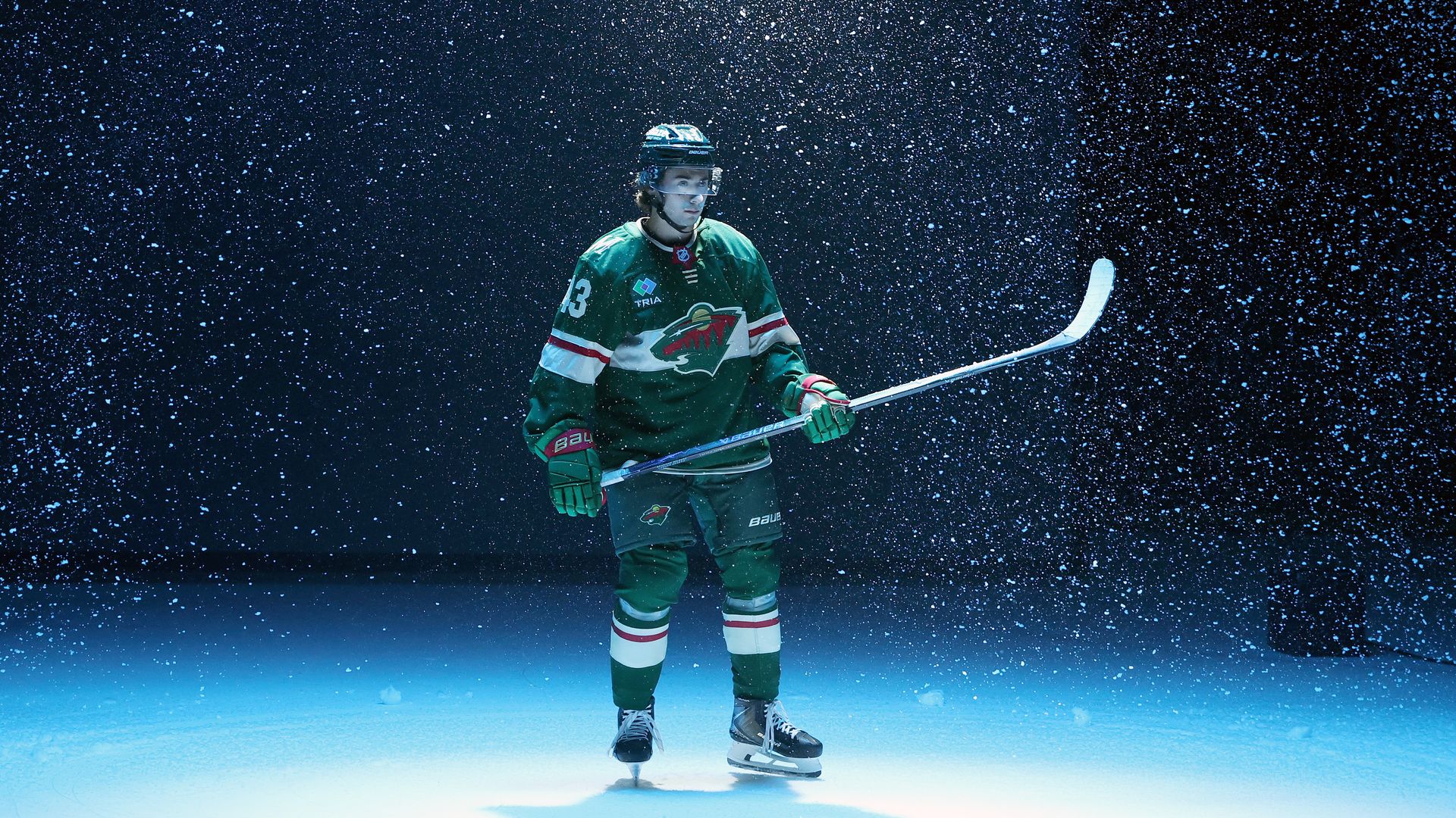 A hockey player in a green jersey stands on an ice rink as snow-like particles fall around a dark blue background, wearing a helmet and gloves while holding a white hockey stick, focused.