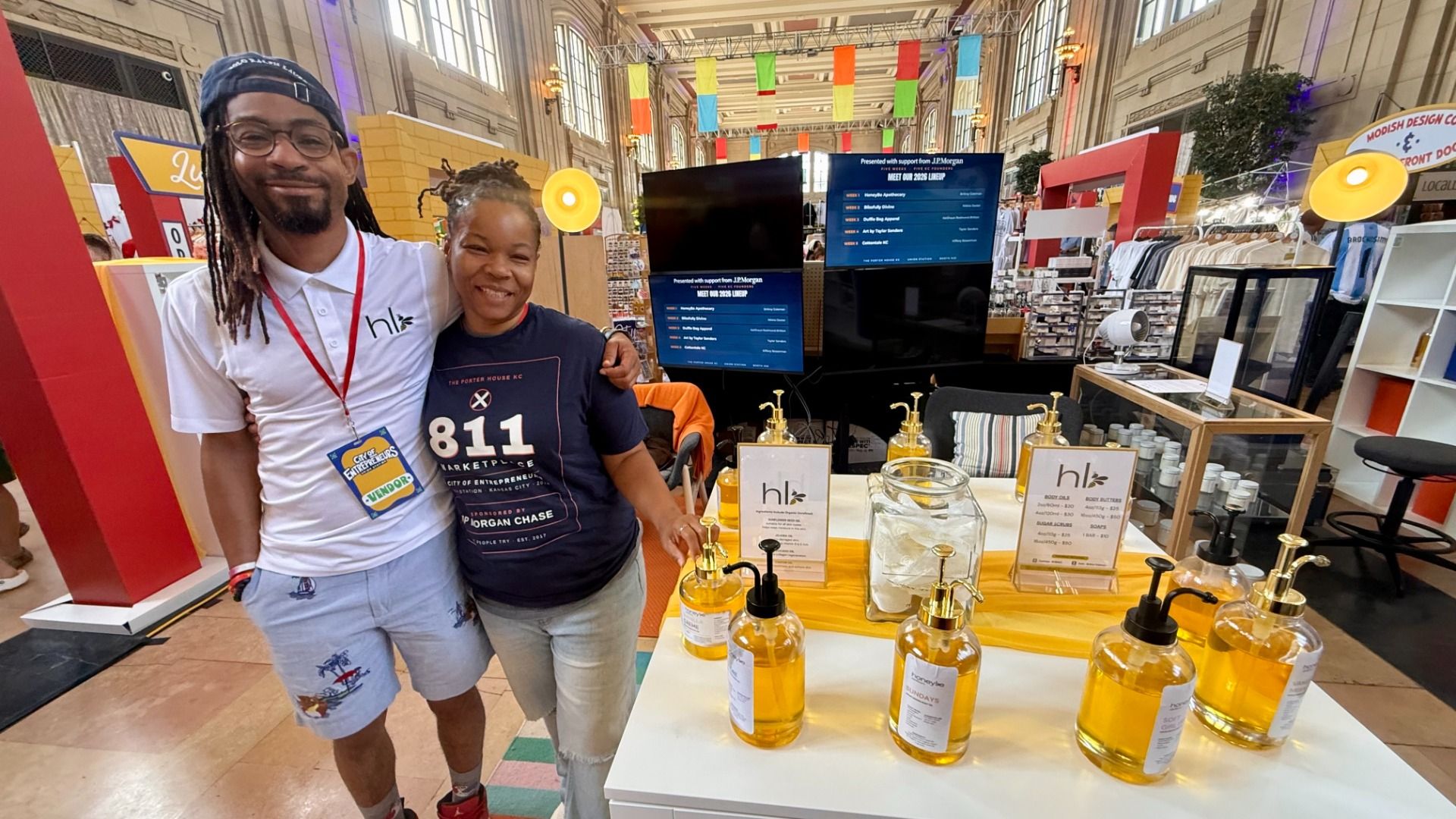 Two smiling vendors pose at a bright trade show booth with yellow liquid bottles on a counter; man in a white polo with hl logo, woman in a navy shirt reading "811 Marketplace"; expo displays and clothing racks behind.