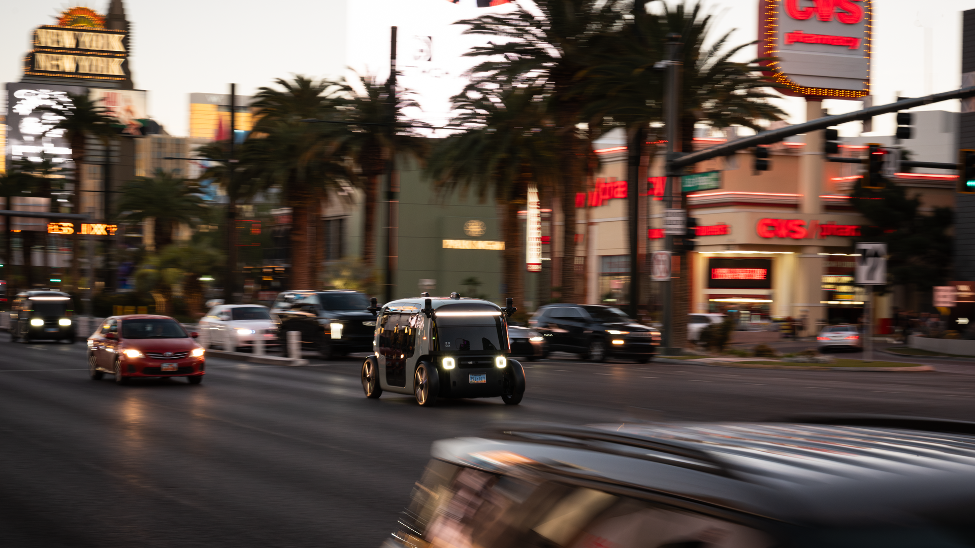 Blurry night street scene on the Las Vegas Strip with palm trees, neon signage including "New York-New York", and a Zoox robotaxi out in front of moving traffic.