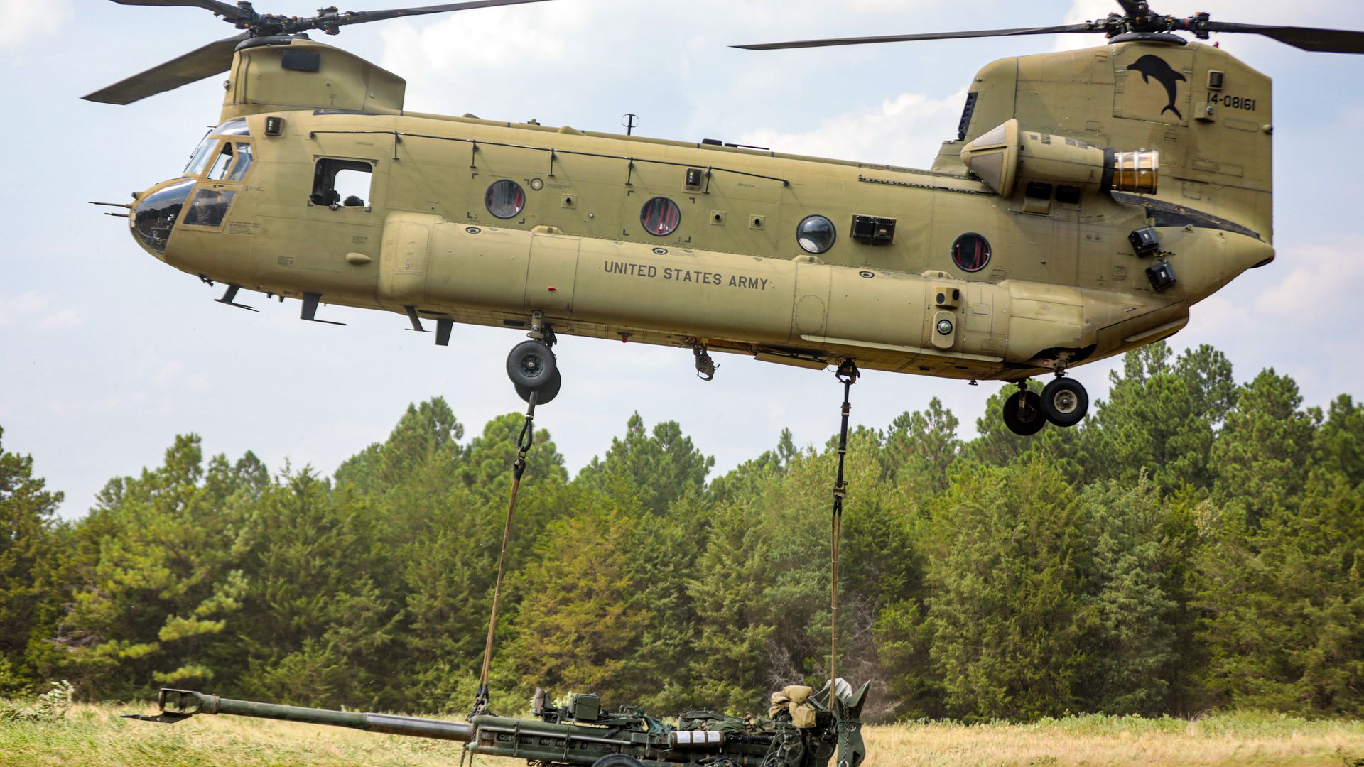 A tan U.S. Army Chinook helicopter airlifts a large green artillery gun over a grassy field with dense green trees in the background under a partly cloudy sky.