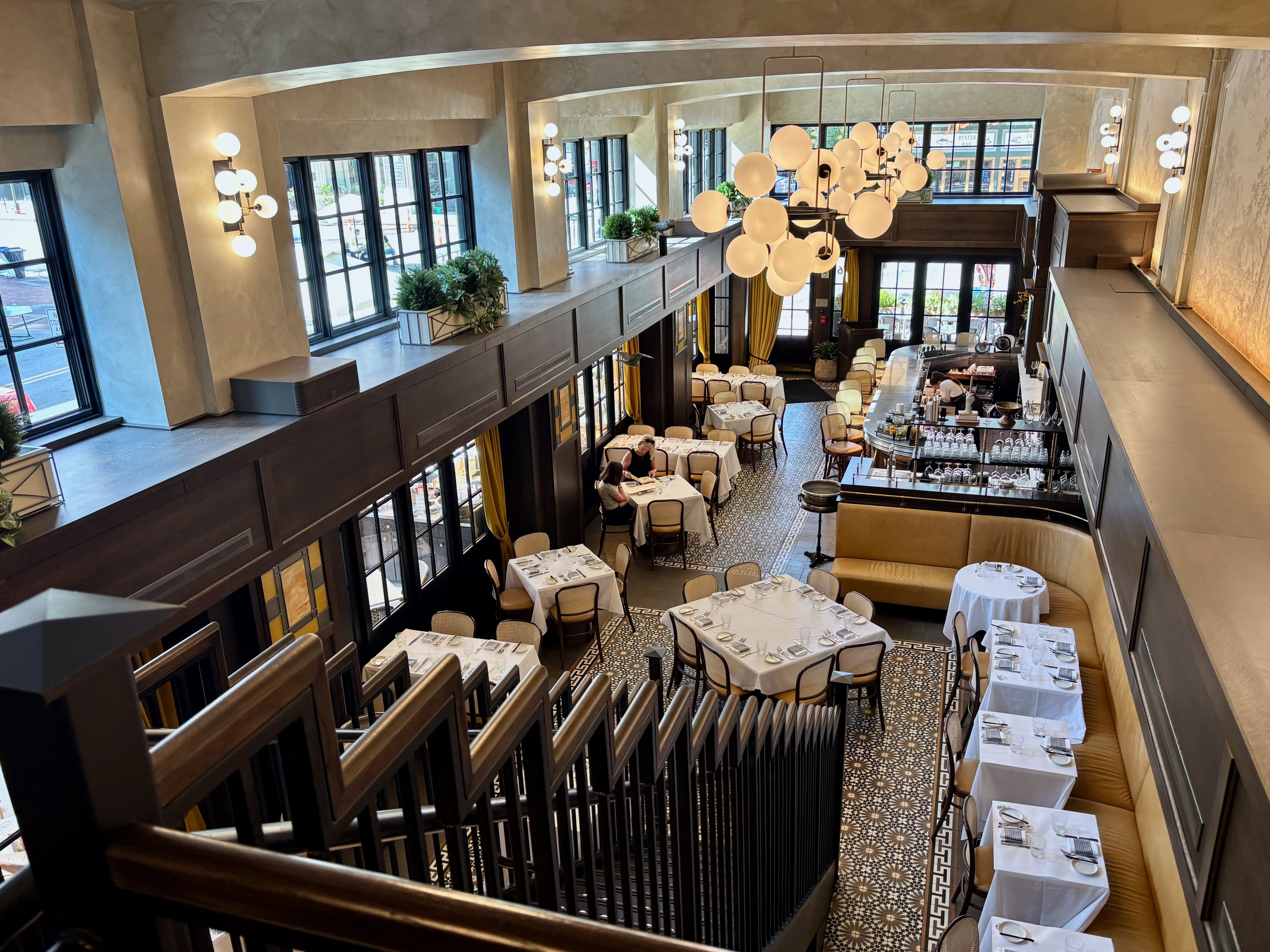 View from a staircase overlooking a bright, elegant restaurant interior with large windows, beige walls, patterned tile floor, white-tablecloth tables, mustard banquette seating, and round globe chandeliers.