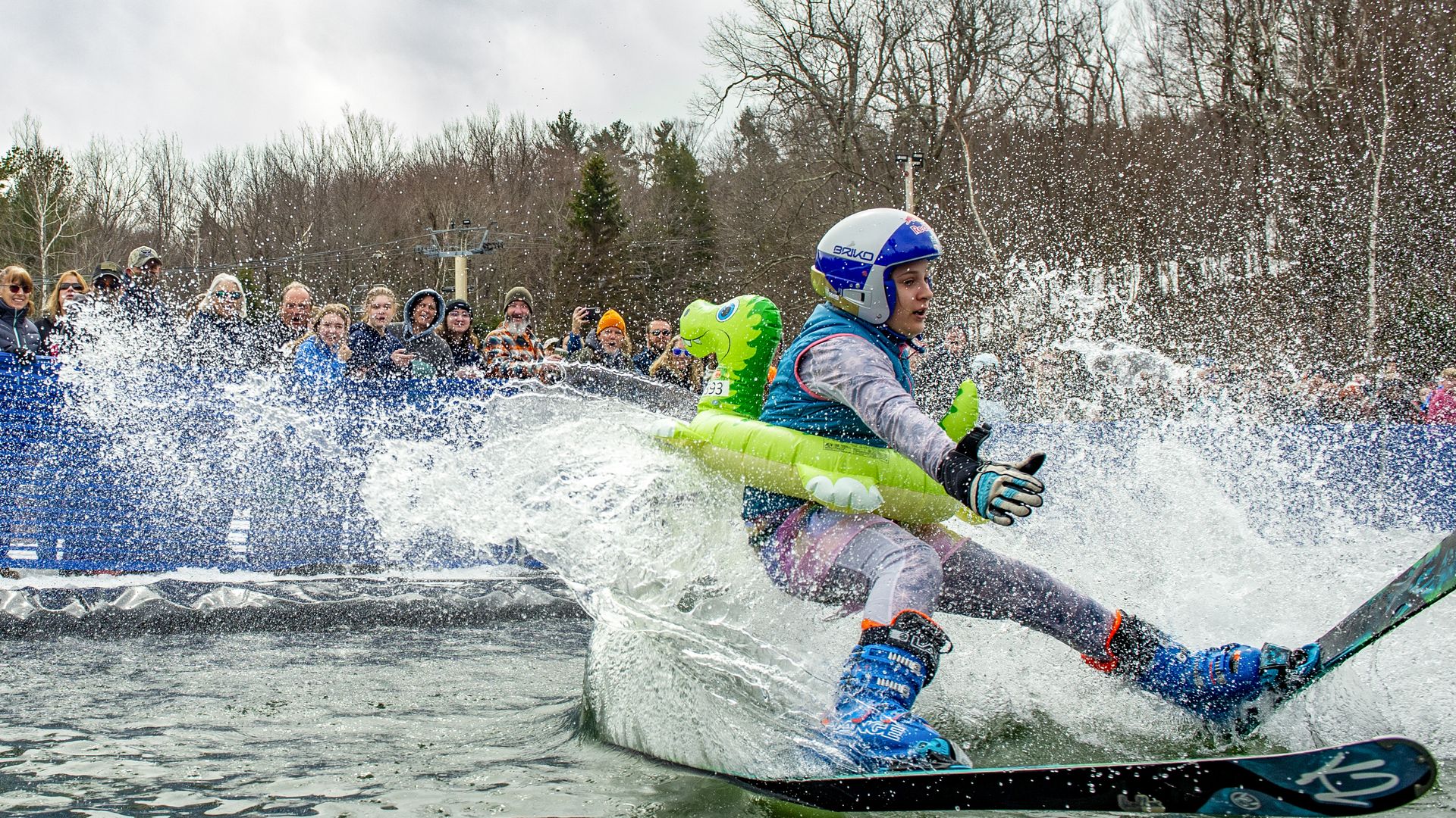 A person on a snowboard attempting to snowboard across a frozen lake. She is dressed in full ski gear and wearing a dinosaur float ring around her chest. A crowd of people watch in the background. 