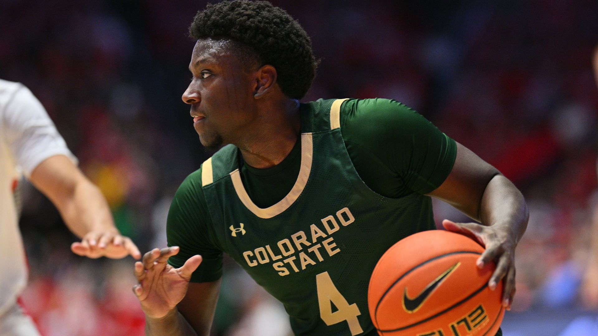 A man in a dark green shirt with the words COLORADO STATE on it and the number four holds a basketball in his left hand during a college basketball game. 