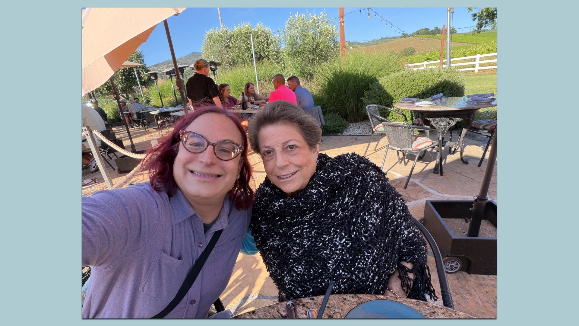 Two smiling women take a selfie at an outdoor patio cafe. In the background, diners sit at tables under beige umbrellas with string lights, against a sunny hillside and white fence.