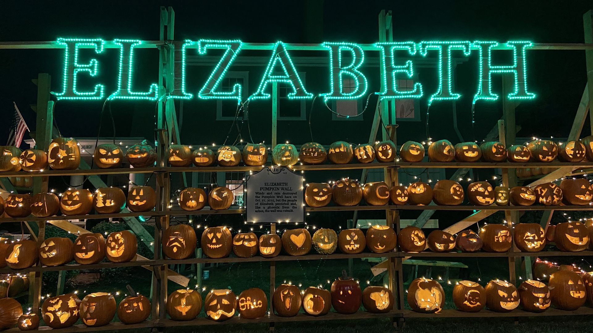Great Elizabeth Pumpkin Wall. Night scene of a pumpkin display on wooden shelves, with carved jack-o'-lanterns and string lights, beneath a bright green neon sign that reads "ELIZABETH" across the top.