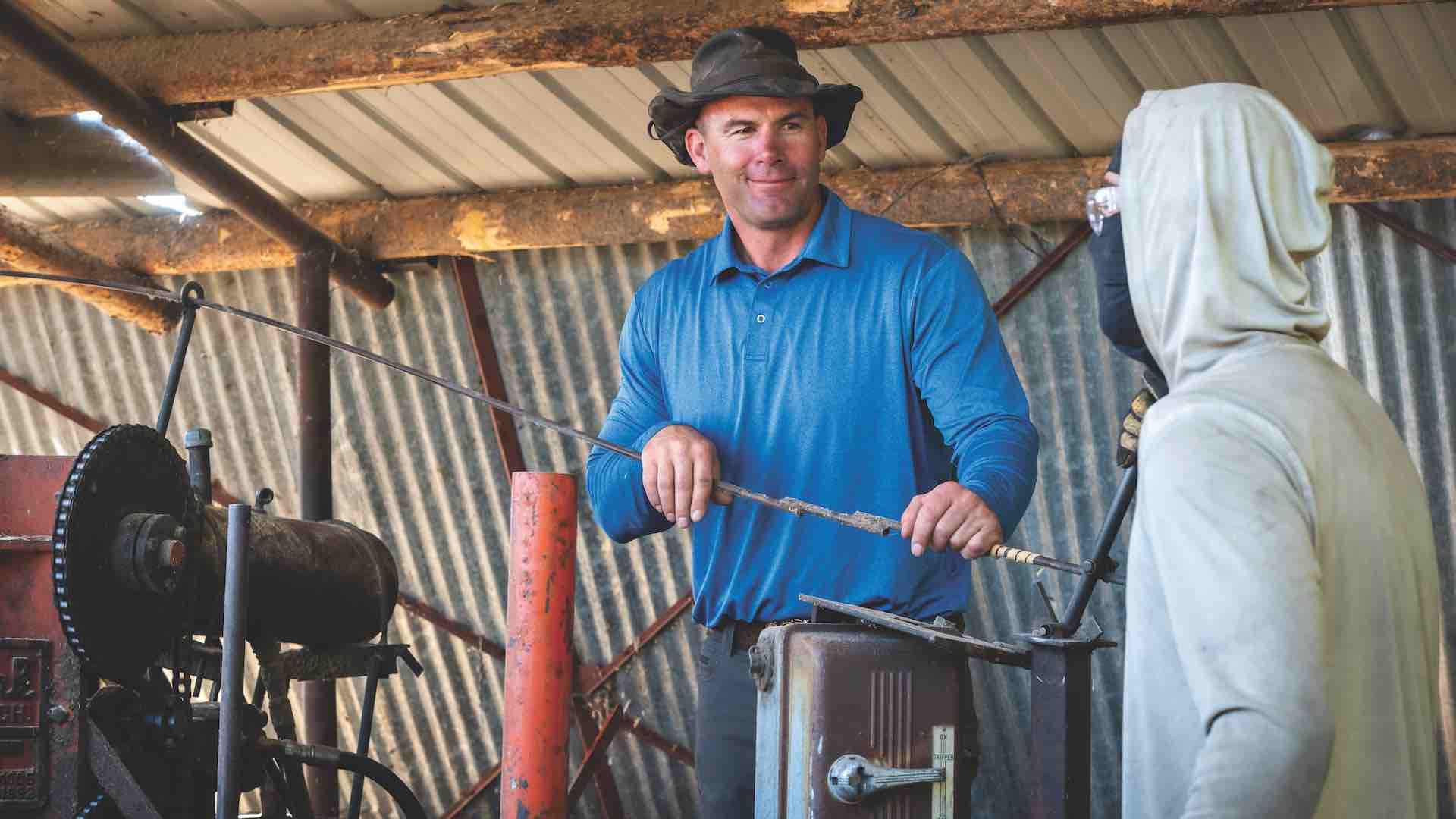 Two men working in a rustic metal shed; one man in a blue shirt and black hat operates a machine while the other in a gray hoodie holds a tool near the machine.