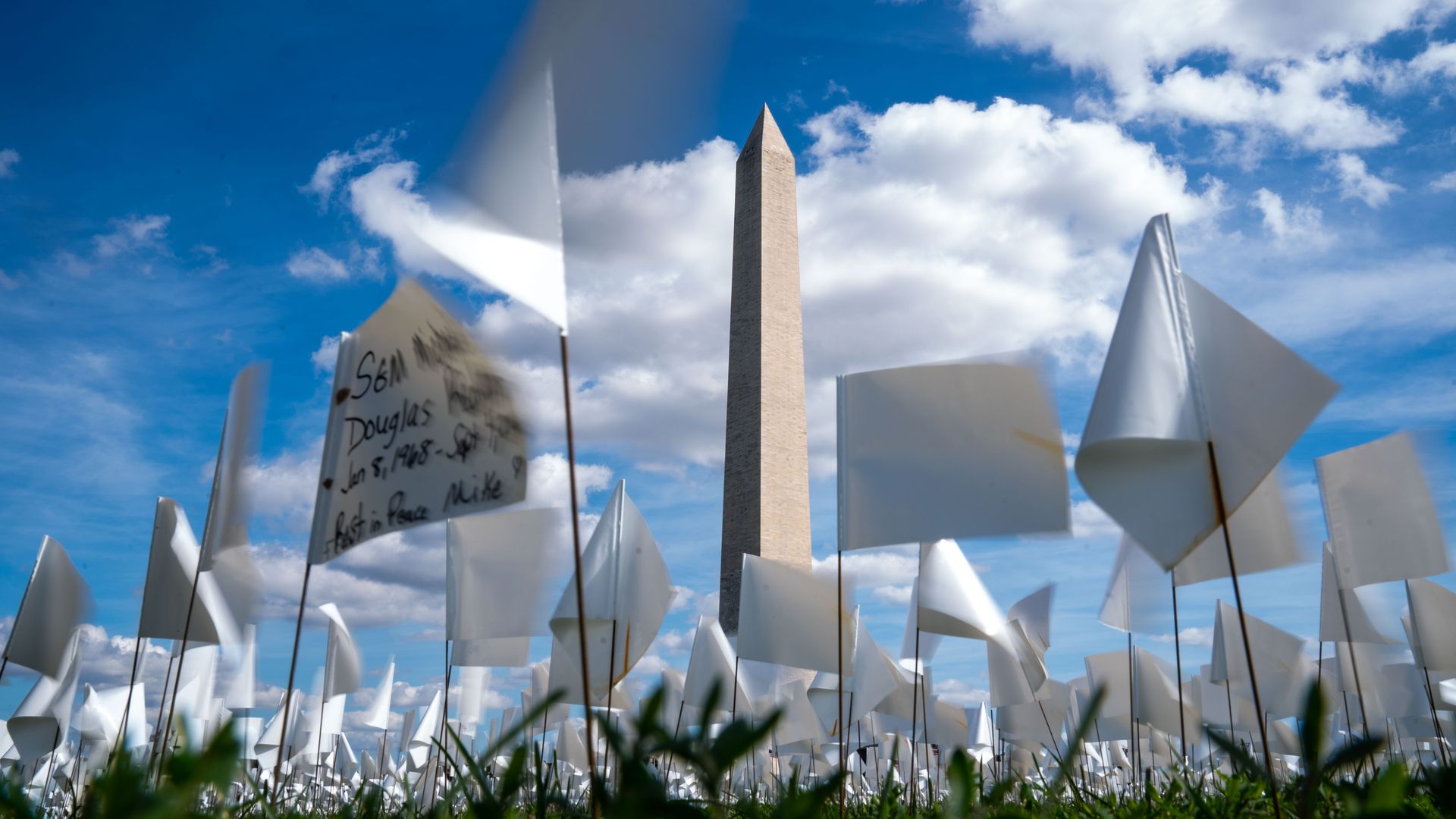 Thousands of mini white flags (dozens in view) are planted in the National Mall, with the Washington Monument in the background.