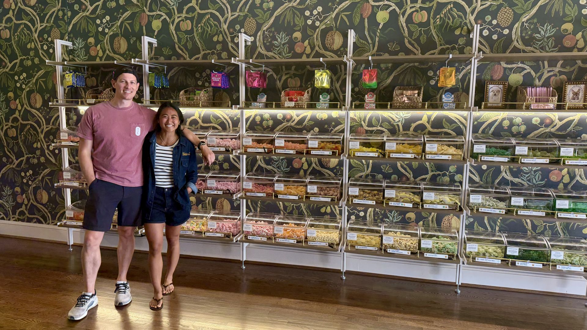 Two friends smile in a candy shop, standing beside glass-fronted display cases filled with colorful sweets; lush green botanical wallpaper featuring vines and fruit covers the wall behind them.