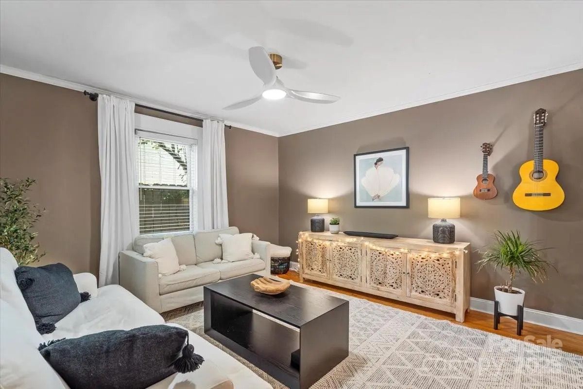 Living room with beige and white sofas, black coffee table, carved wooden sideboard with string lights and lamps, brown walls with two guitars hanging, a plant, and framed artwork.